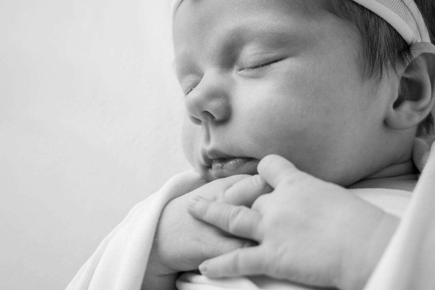 A sleeping newborn baby wearing white clothing rests peacefully against a light background.