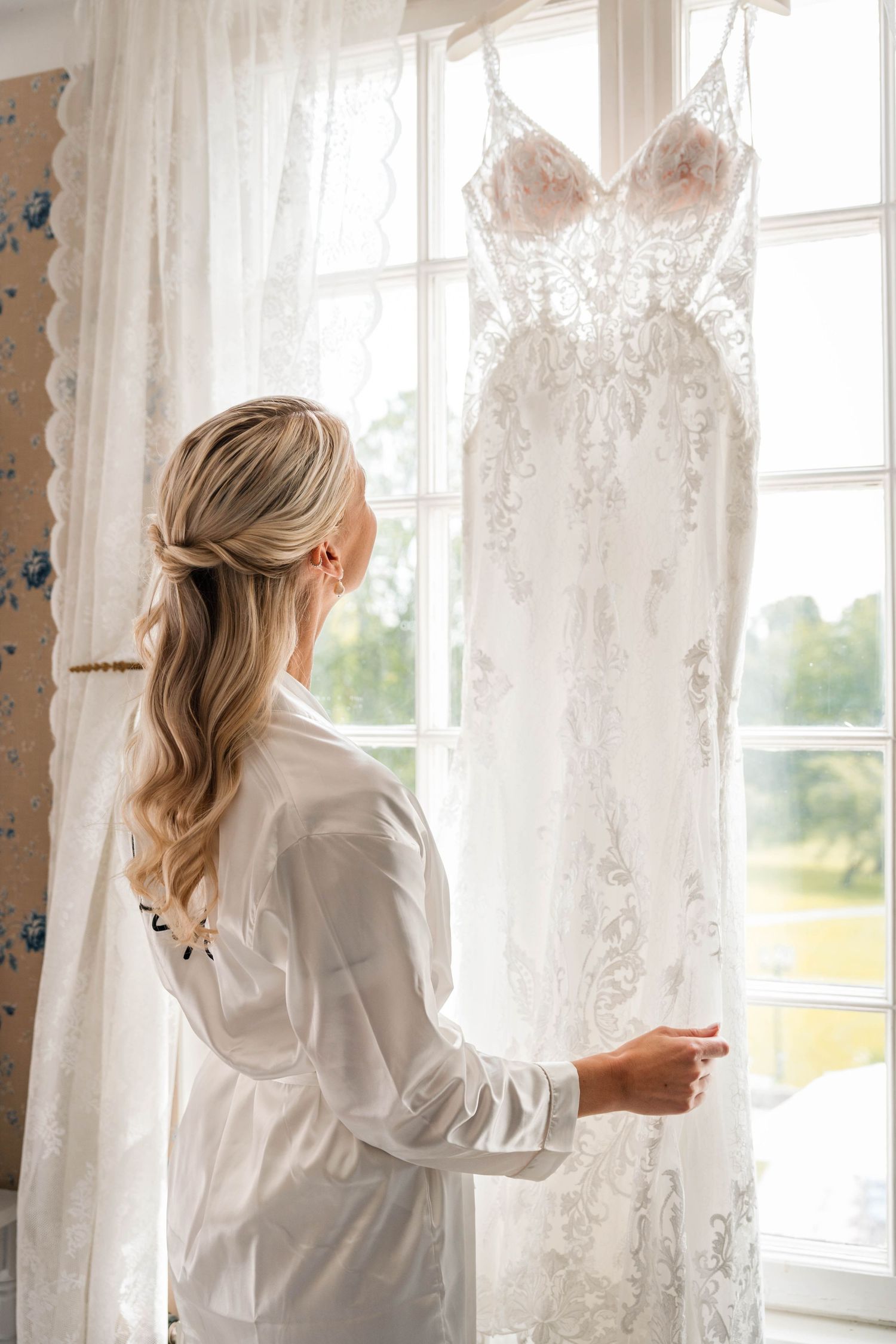 Sequence of a woman in white adjusting delicate lace curtains by a bright window.