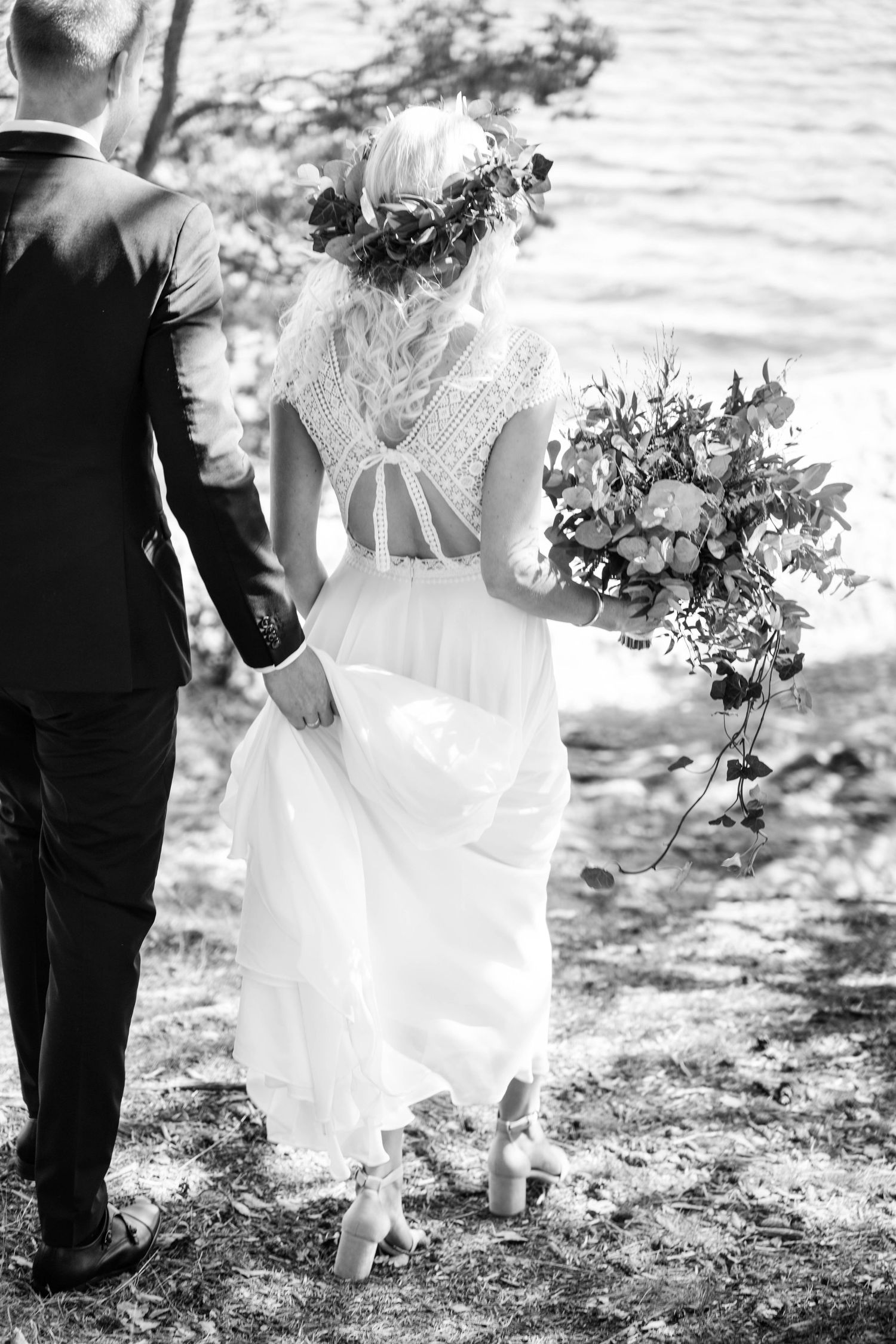 Black and white photo of bride in lace back wedding dress with floral crown and cascading bouquet walking outdoors.