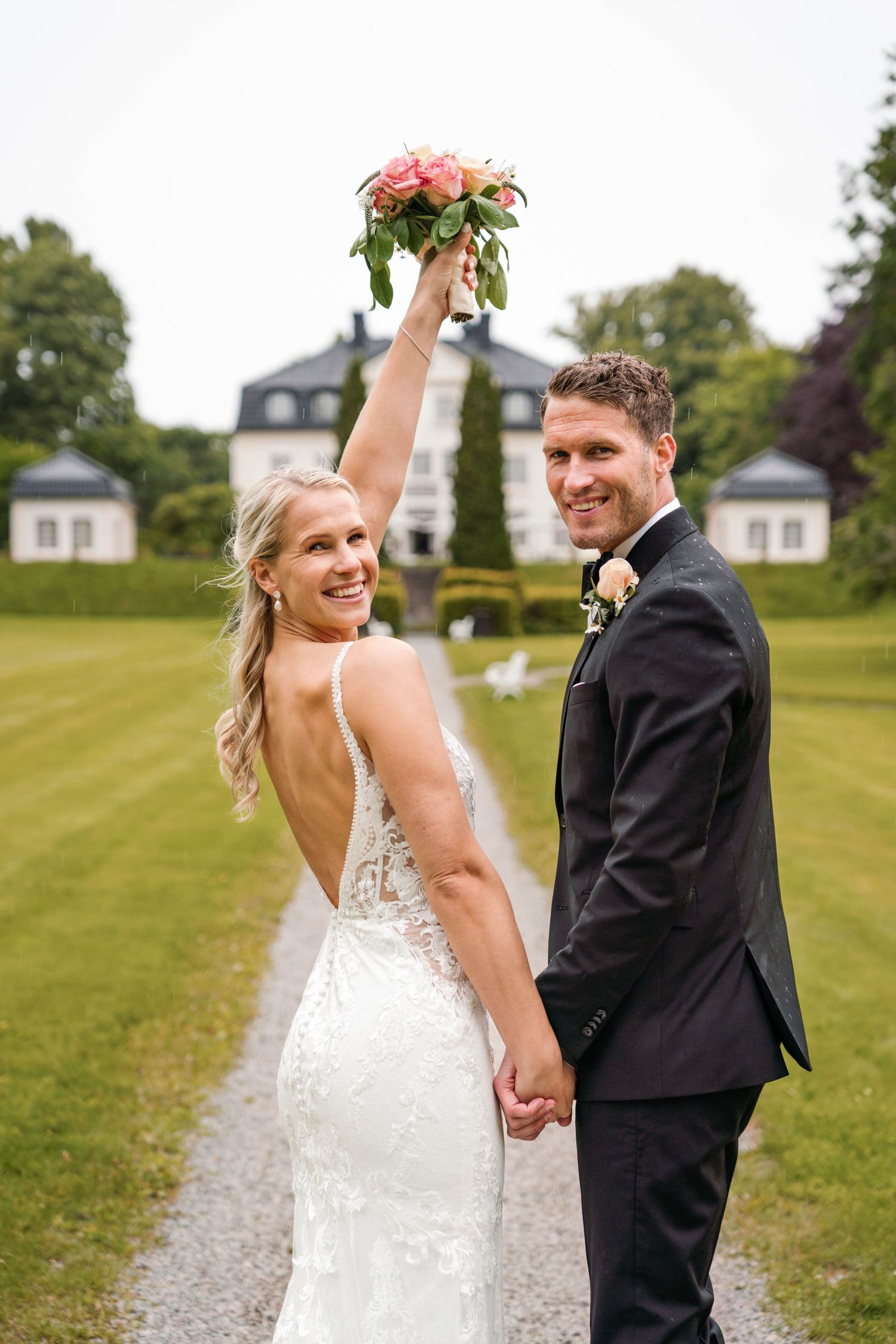 Romantic wedding couple walking together on a gravel path with estate buildings in background.