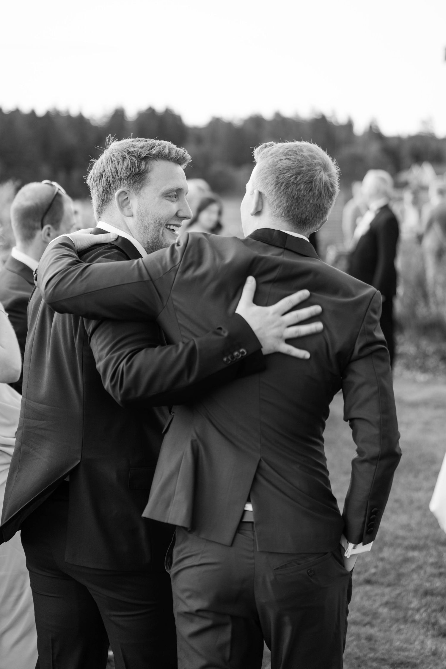 Groomsmen in black suits sharing emotional group hugs at outdoor wedding celebration.