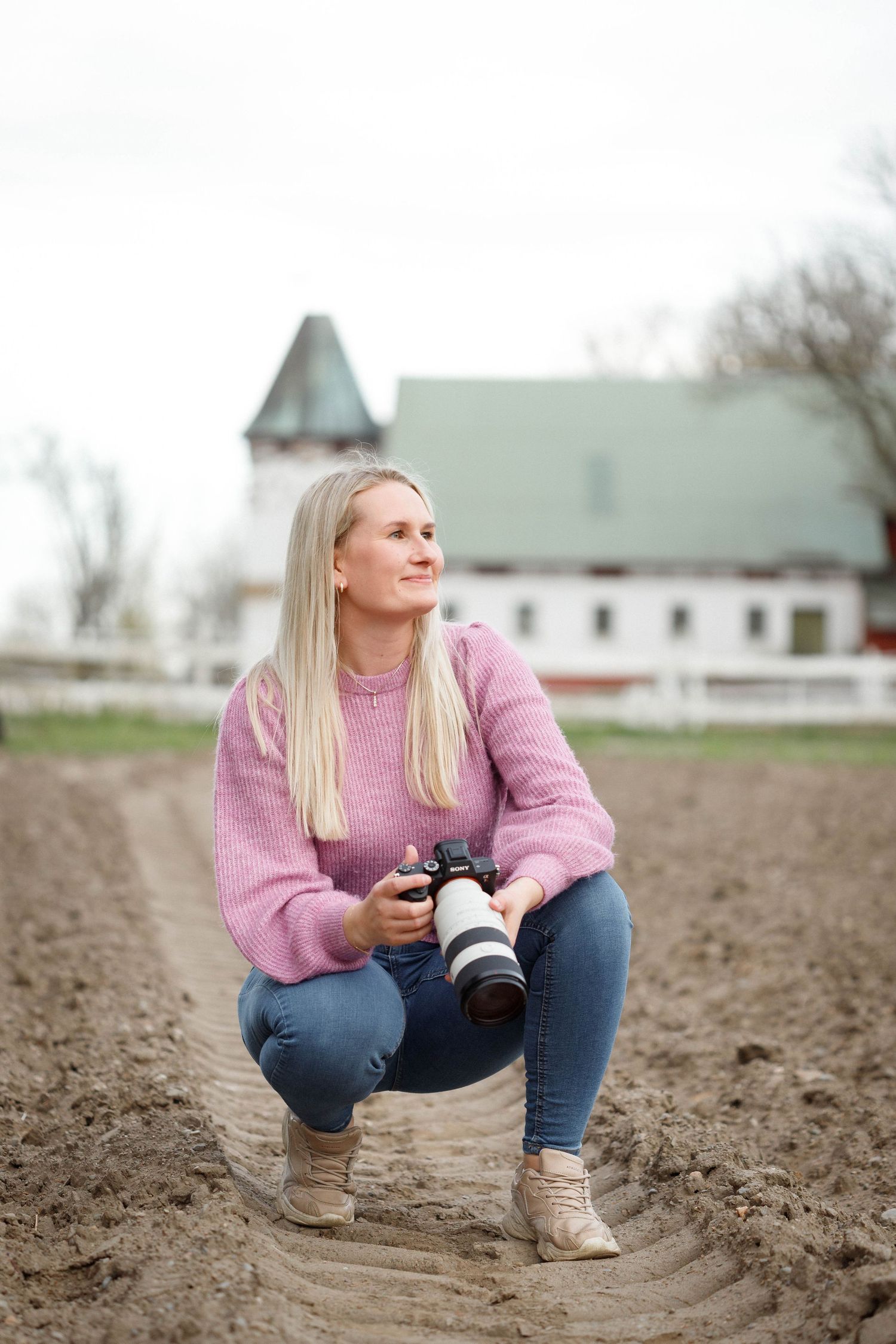 Photographer in pink sweater and jeans crouches in a plowed field with camera near rural buildings.