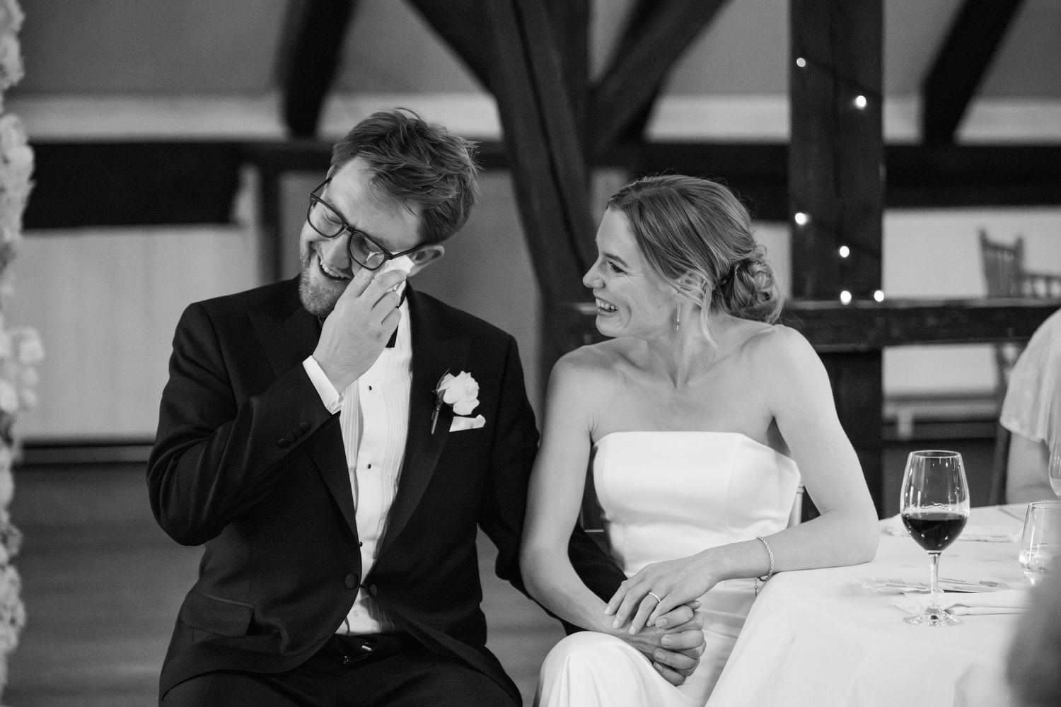 Black and white wedding reception photo showing guests laughing and enjoying wine at an elegant dinner table.