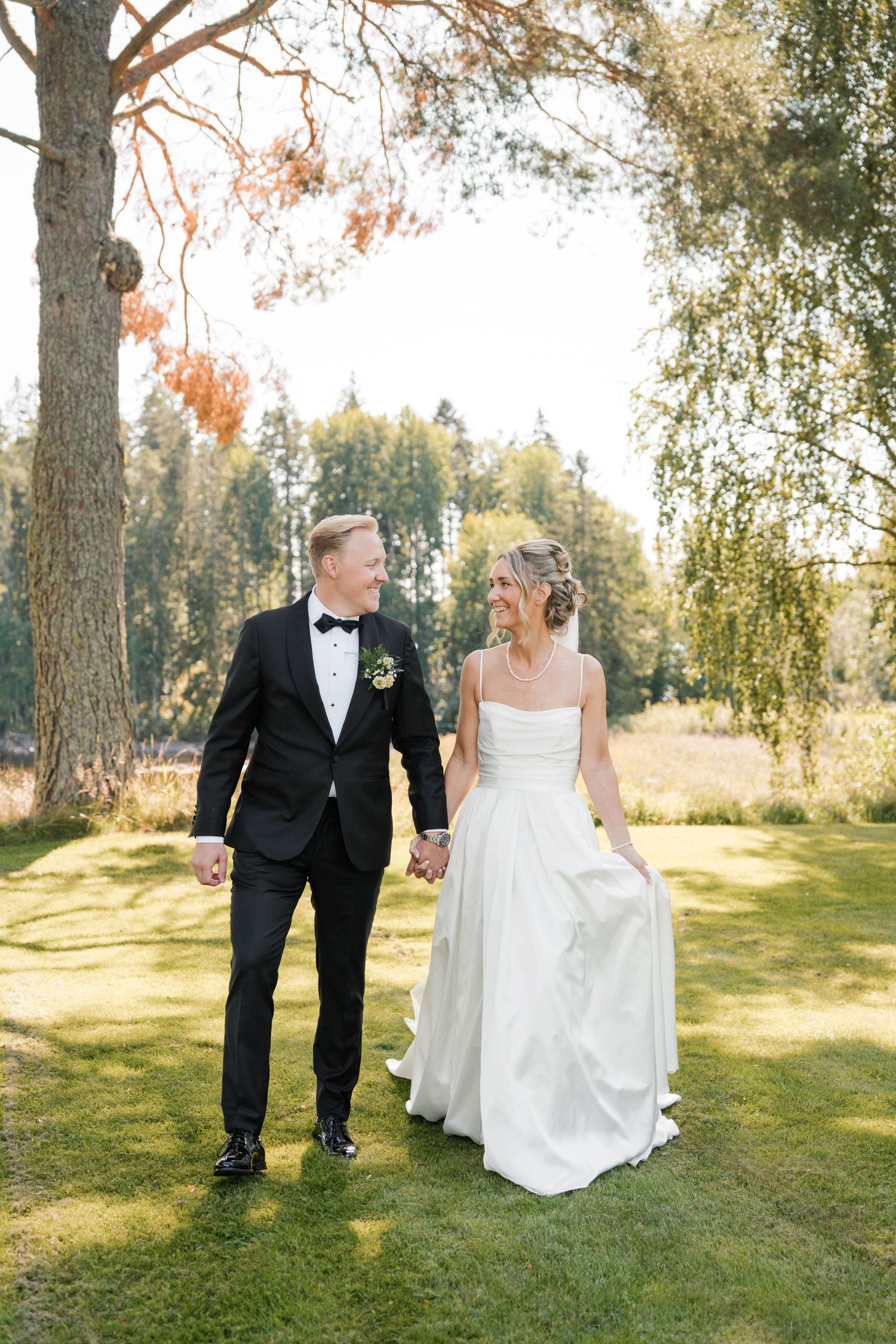 Newlyweds walk hand in hand through sunlit garden with birch trees.