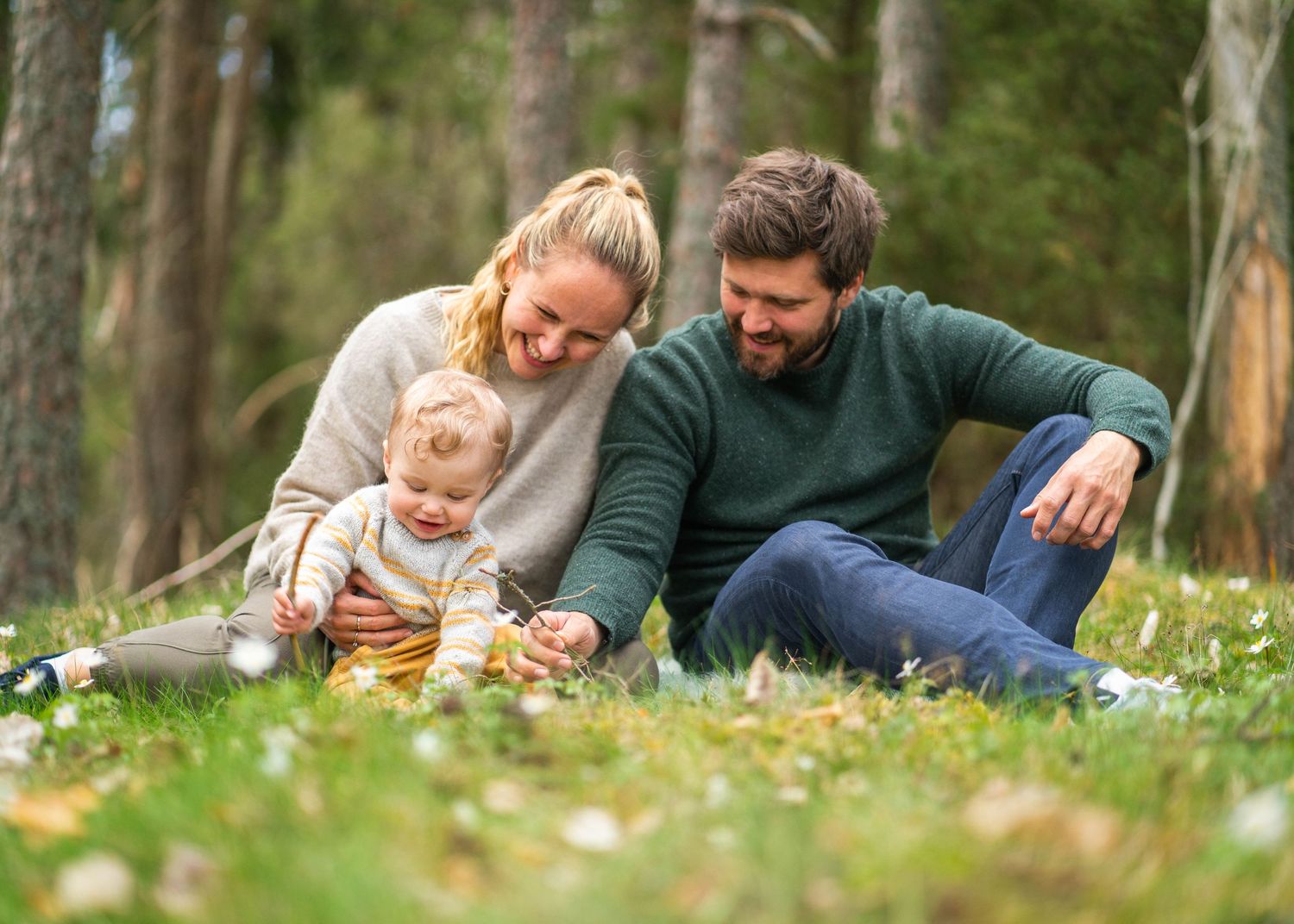 Family relaxing together on grass in a wooded area with wildflowers.