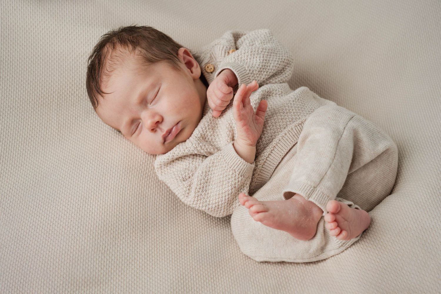 A sleeping newborn baby wrapped in soft beige knit fabric lying on a neutral backdrop.