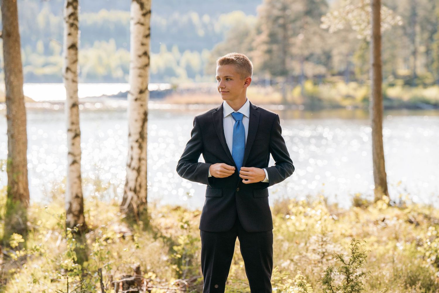 Professional wearing a dark suit stands in a wooded outdoor setting with sunlit trees and water in the background.
