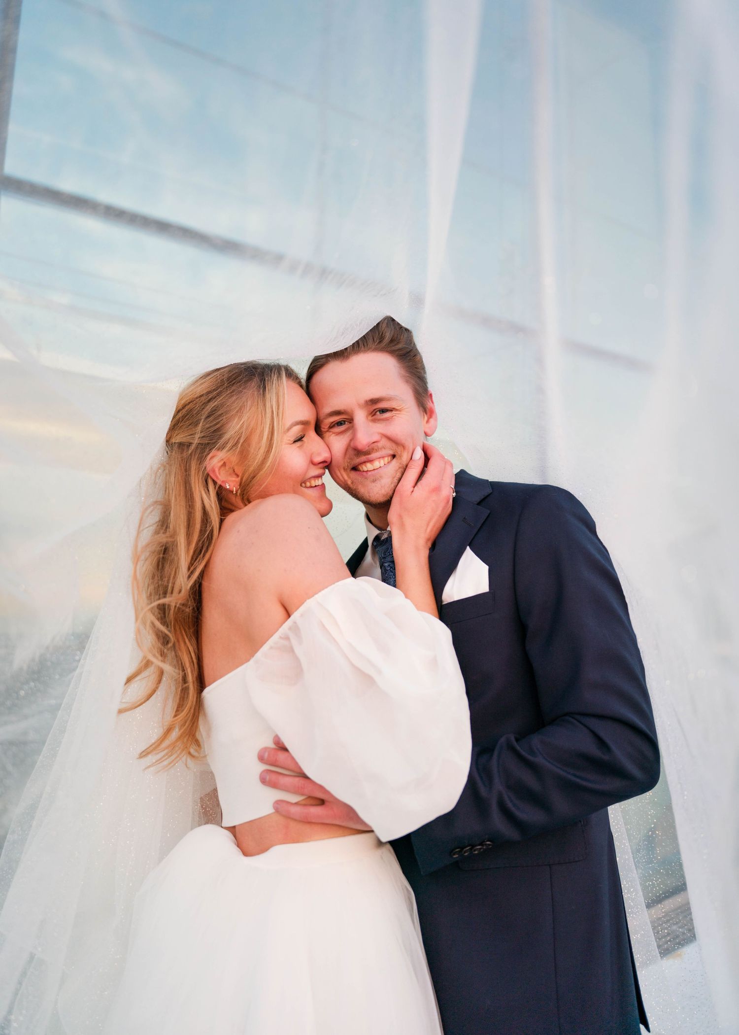 Romantic wedding portrait of couple embracing against sunset sky with sheer white curtains.