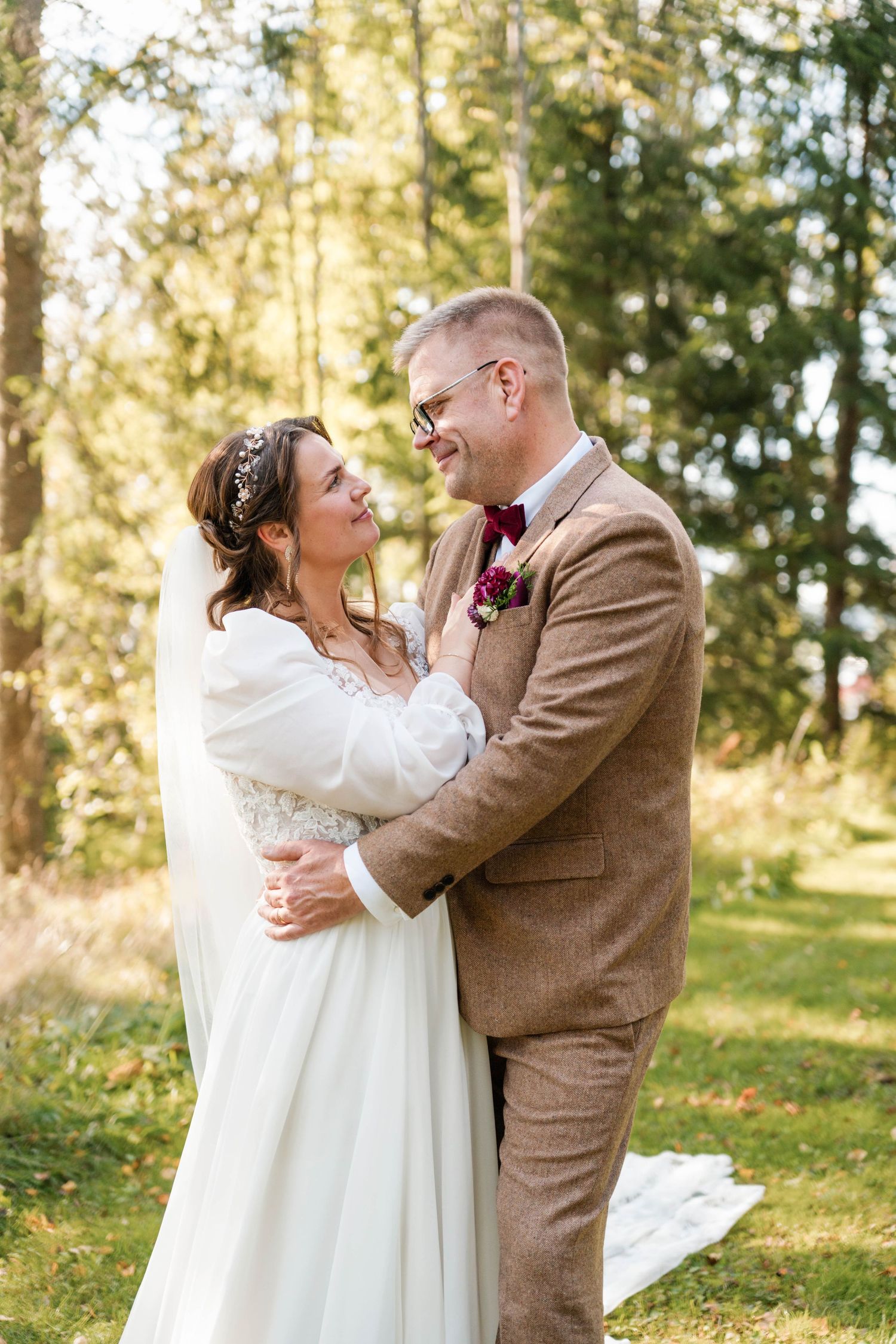 Wedding couple embraces in sunlit forest wearing white dress and brown suit.