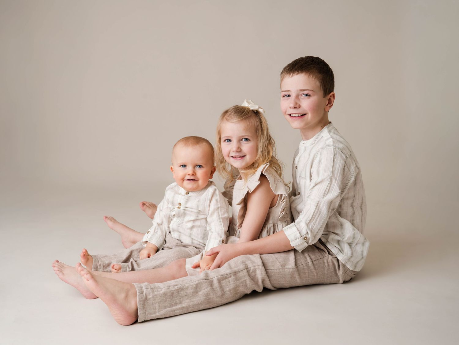 Three children pose together on a neutral background, wearing coordinating cream and beige outfits in a professional studio portrait.