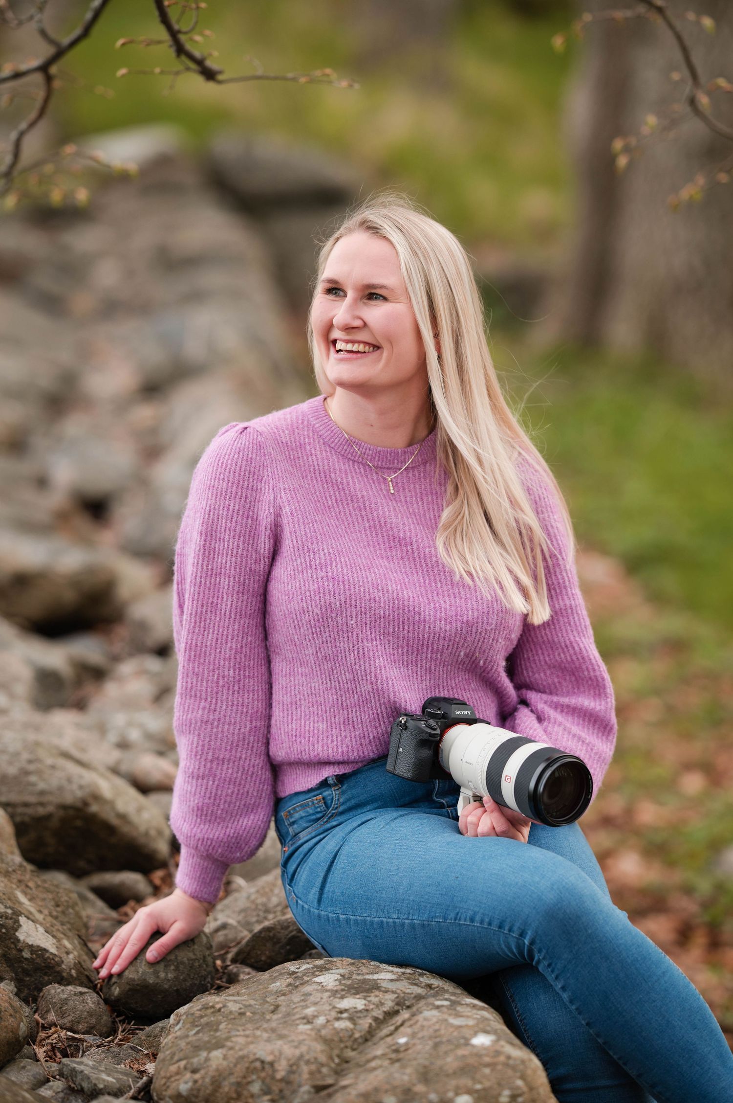 Photographer wearing lilac sweater sits on rocks while holding professional camera equipment.