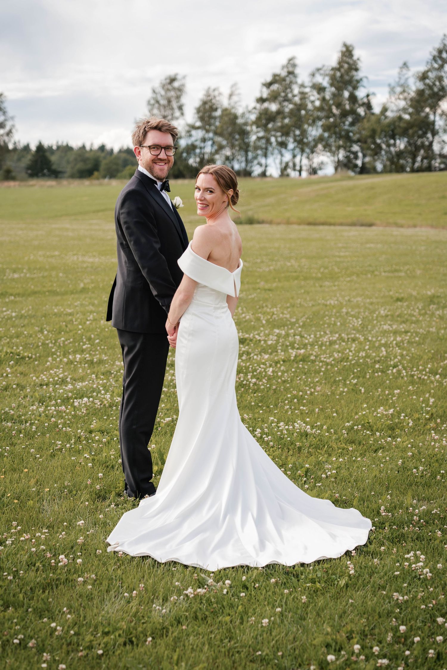 A couple in wedding attire stands together in a sunny meadow filled with small white wildflowers.