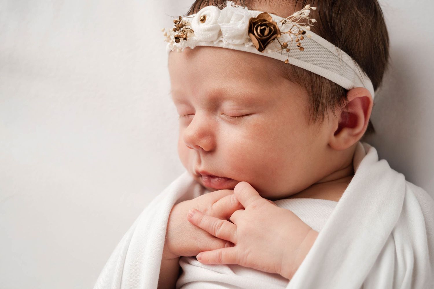 A collage of four photos showing a sleeping newborn baby wearing a white headband and wrapped in white fabric.