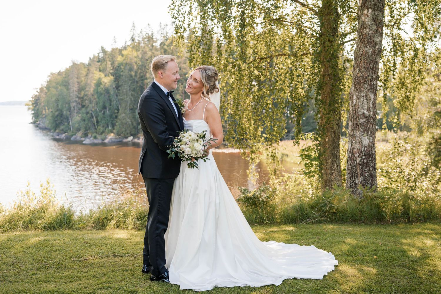 A romantic wedding couple embraces by a serene lake with birch trees and sunlit nature in the background.