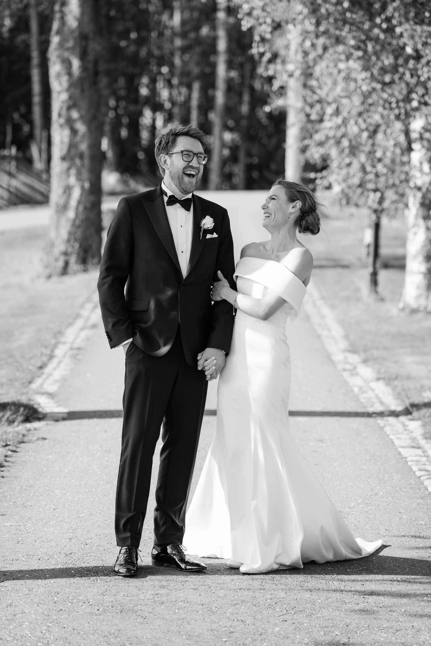 A joyful bride and groom share a candid laughing moment on a tree-lined path in black and white photography.