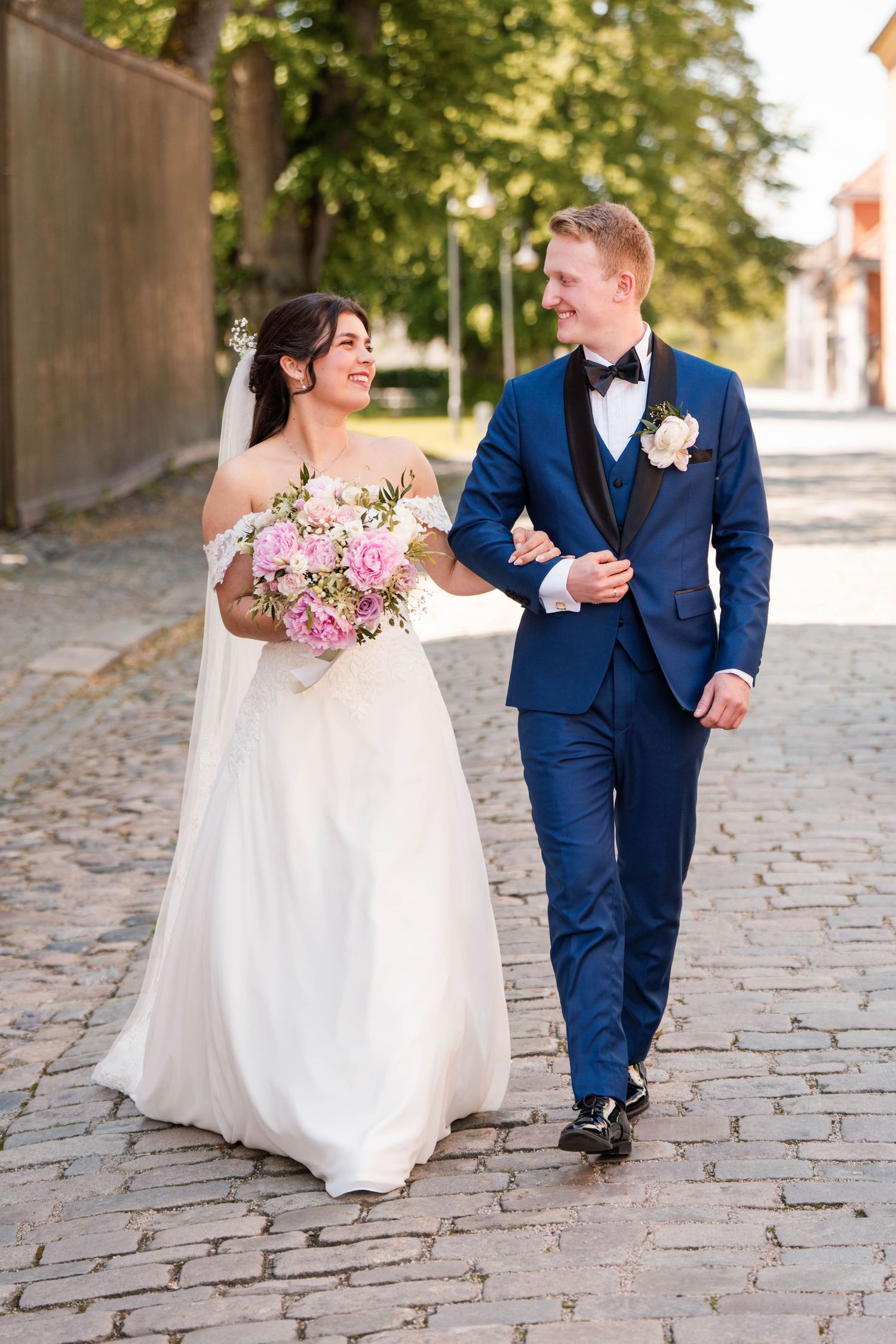 Newlyweds walking together on cobblestone path with pink floral bouquet.