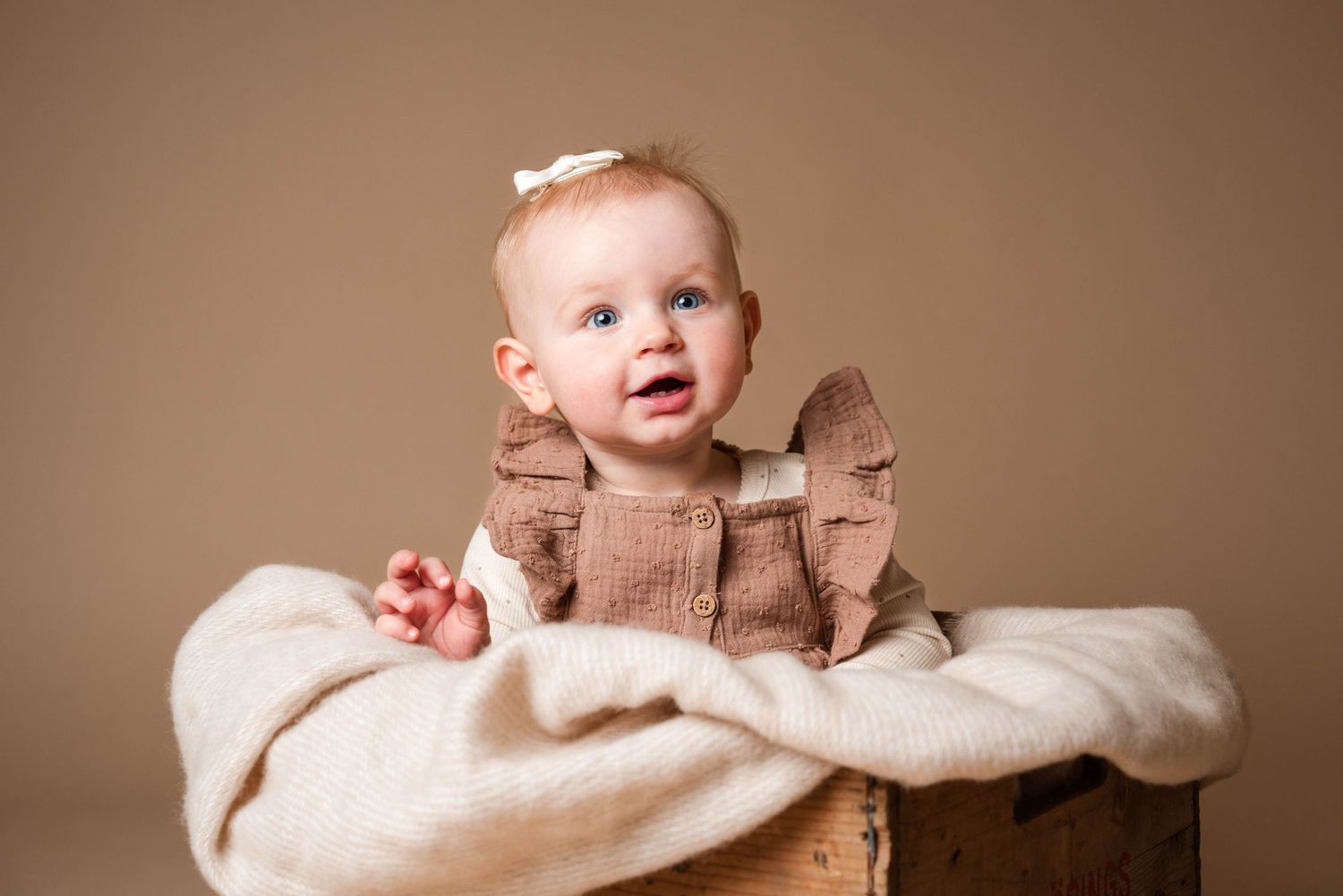 A baby in a brown dress sits on a white blanket with a joyful expression.