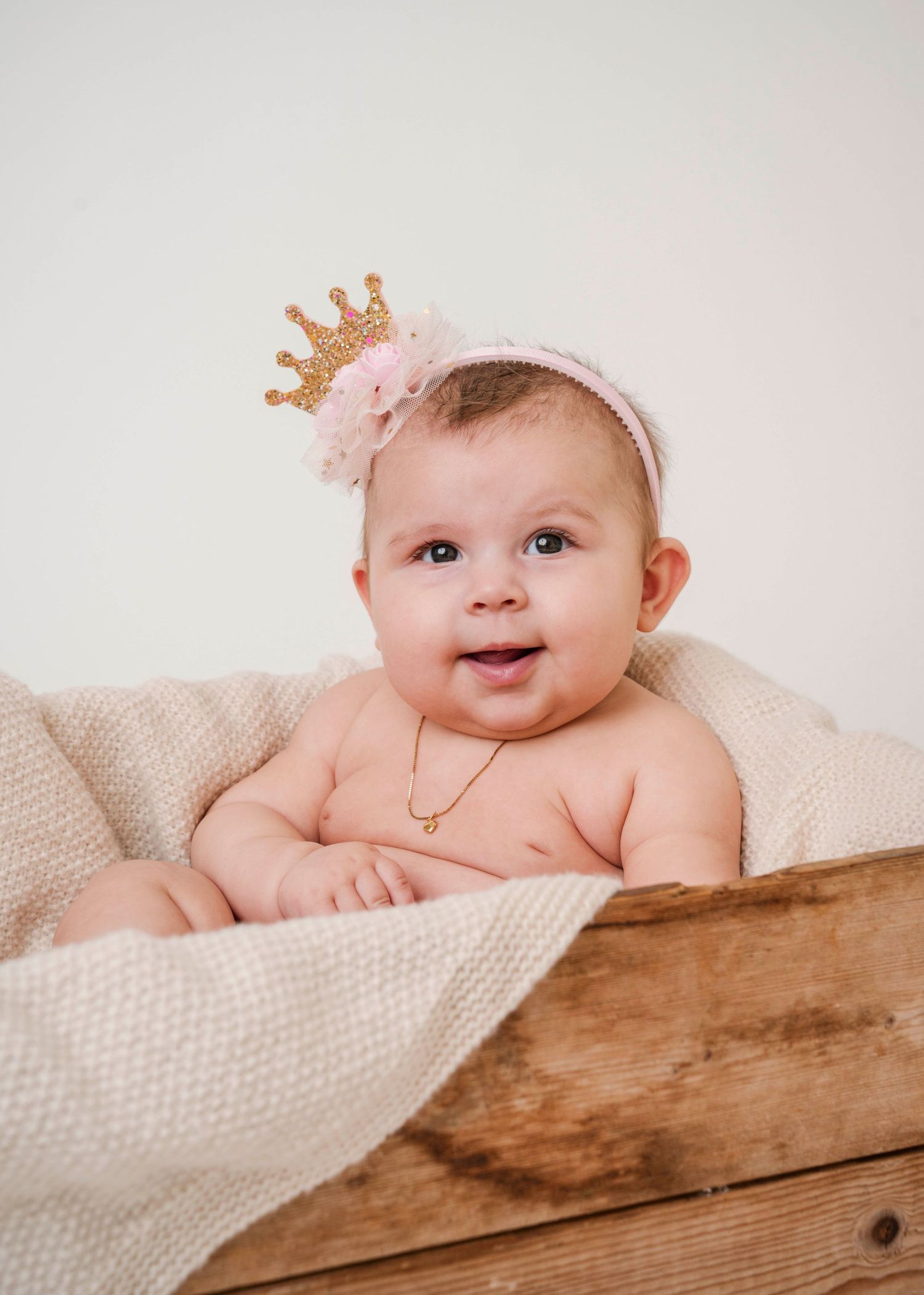 Baby wearing pink crown lying on wooden surface with cream blanket.