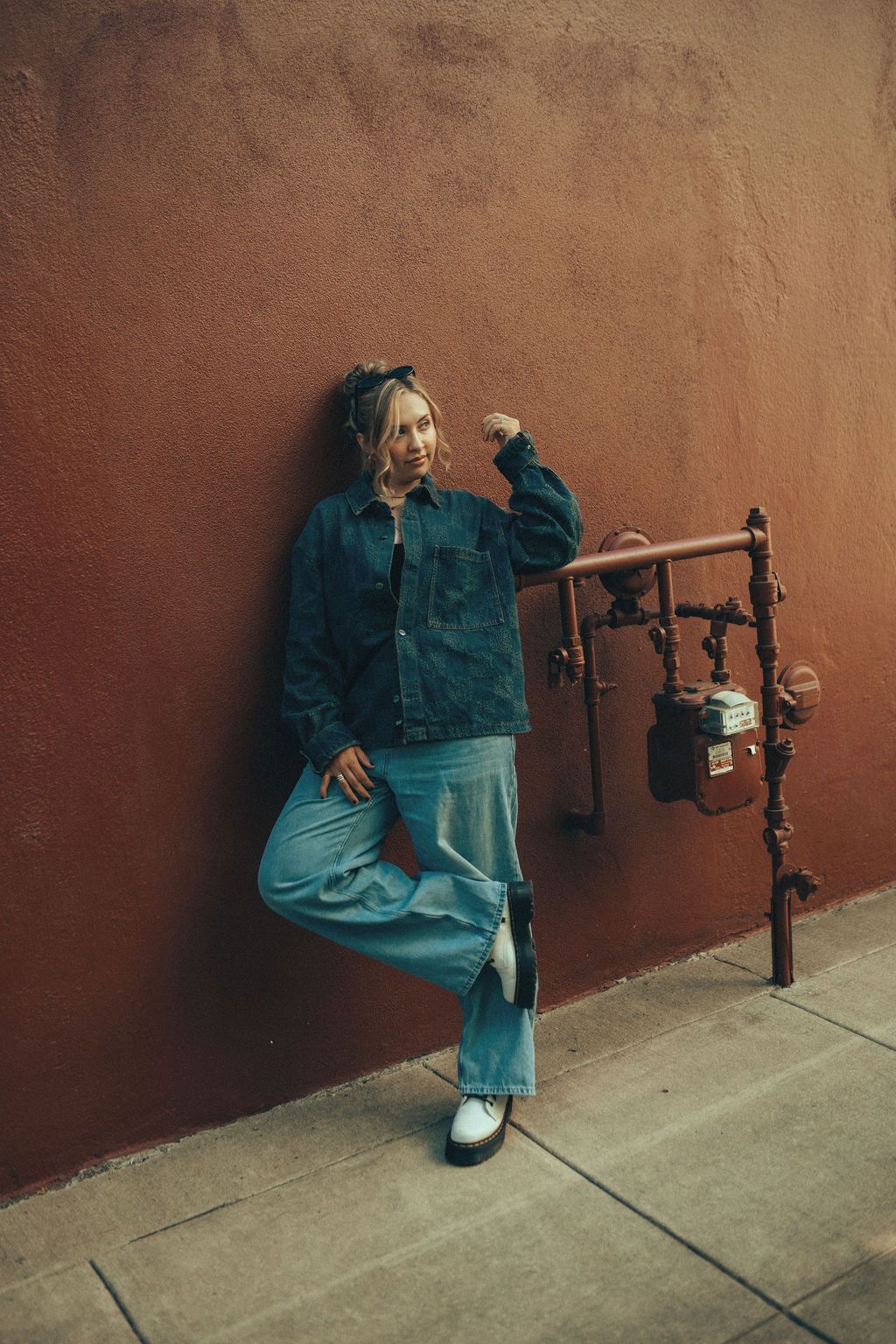 Figure in double denim outfit leaning against rustic brown wall with metal pipes.