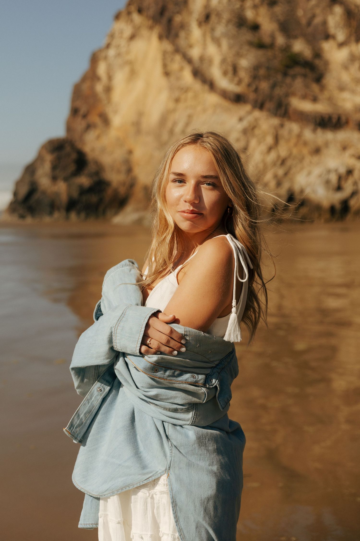 Person in denim jacket posing against rocky coastal backdrop during golden hour.