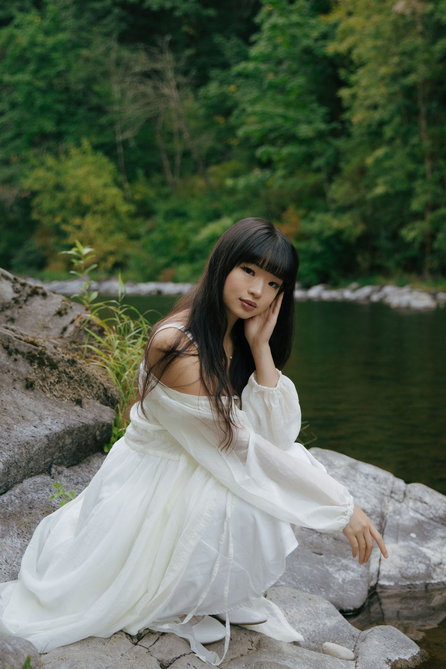 Figure in white flowing dress posing gracefully on rocks by a serene lake.