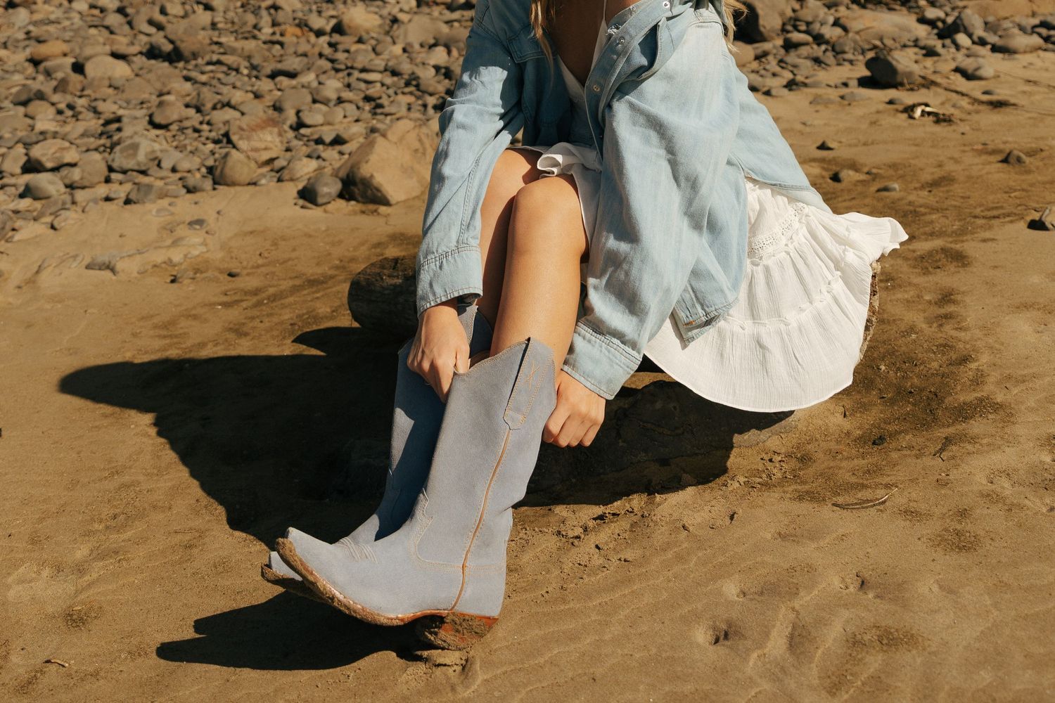Person in white dress and denim walking on sandy beach near cliffs.