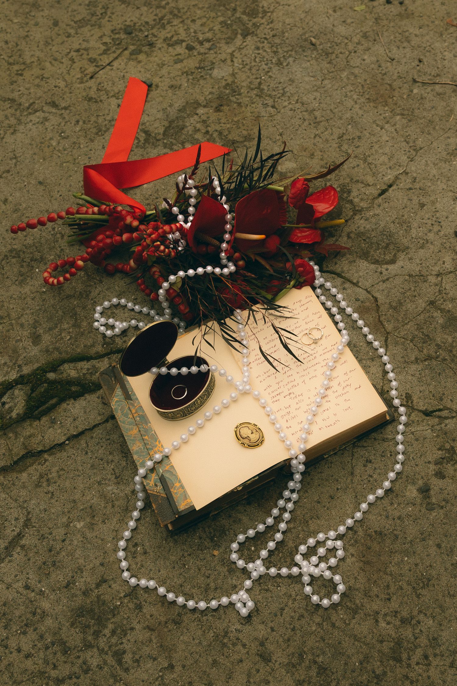 Vintage book with pearl necklace, gold brooch, ring box, and red floral bouquet with ribbon on stone surface.