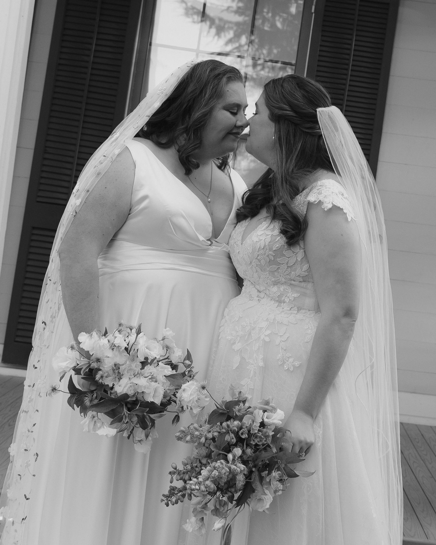 Black and white photo of a bride in a white wedding dress and veil holding a floral bouquet on a porch.