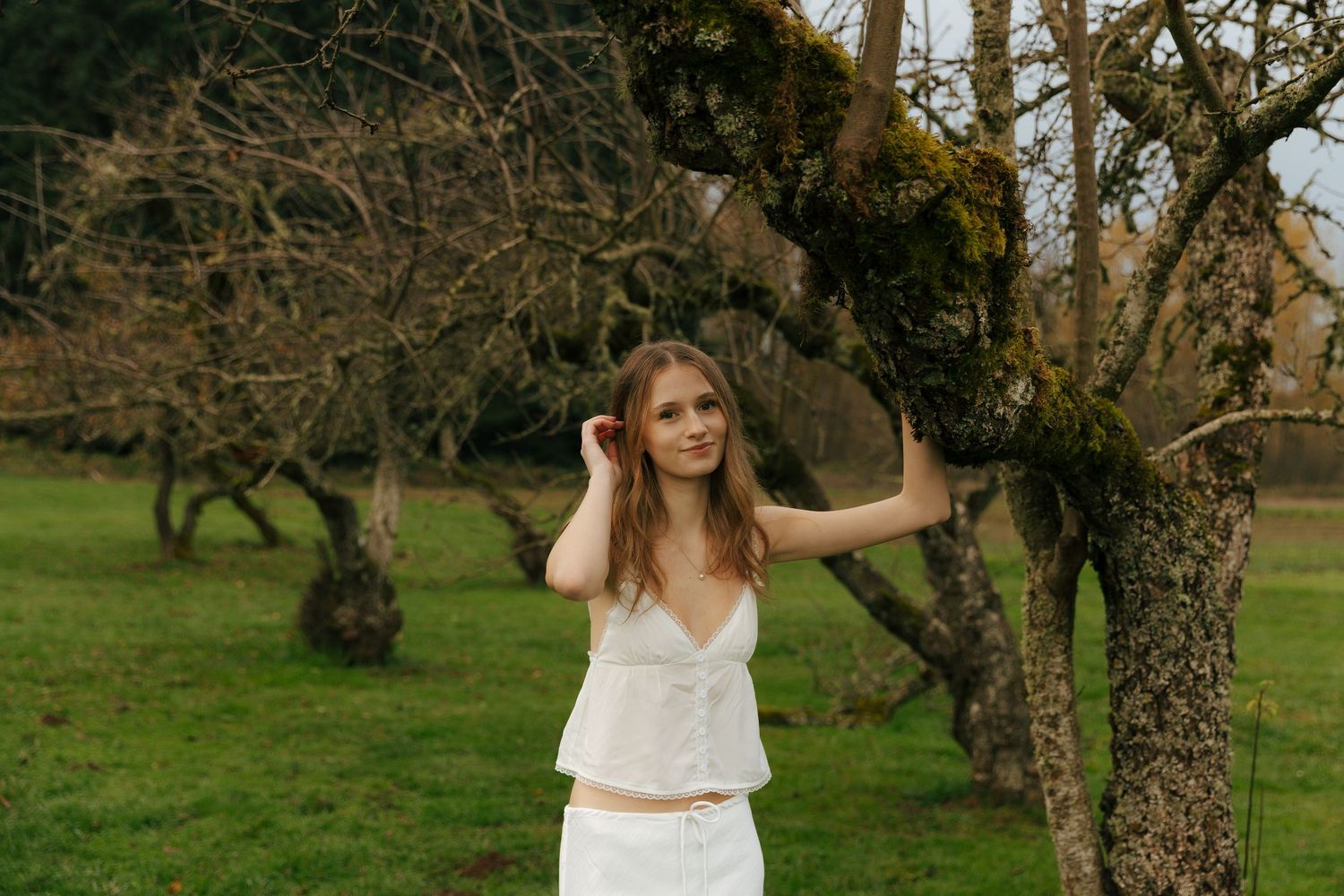 White dress flowing in breeze among bare fruit trees in green field.
