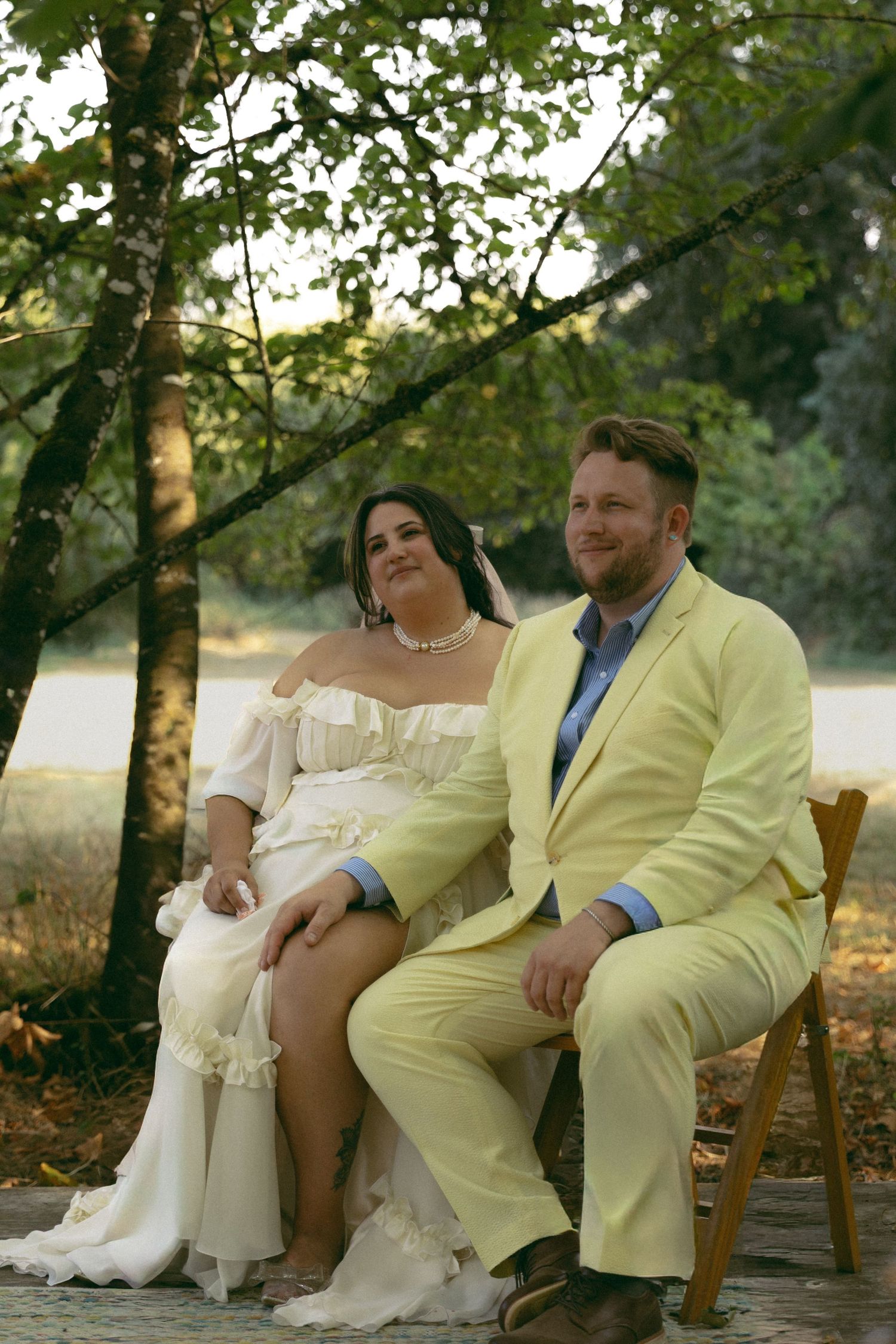 Bride in white ruffled dress and groom in yellow suit sitting together outdoors under trees for wedding portrait.