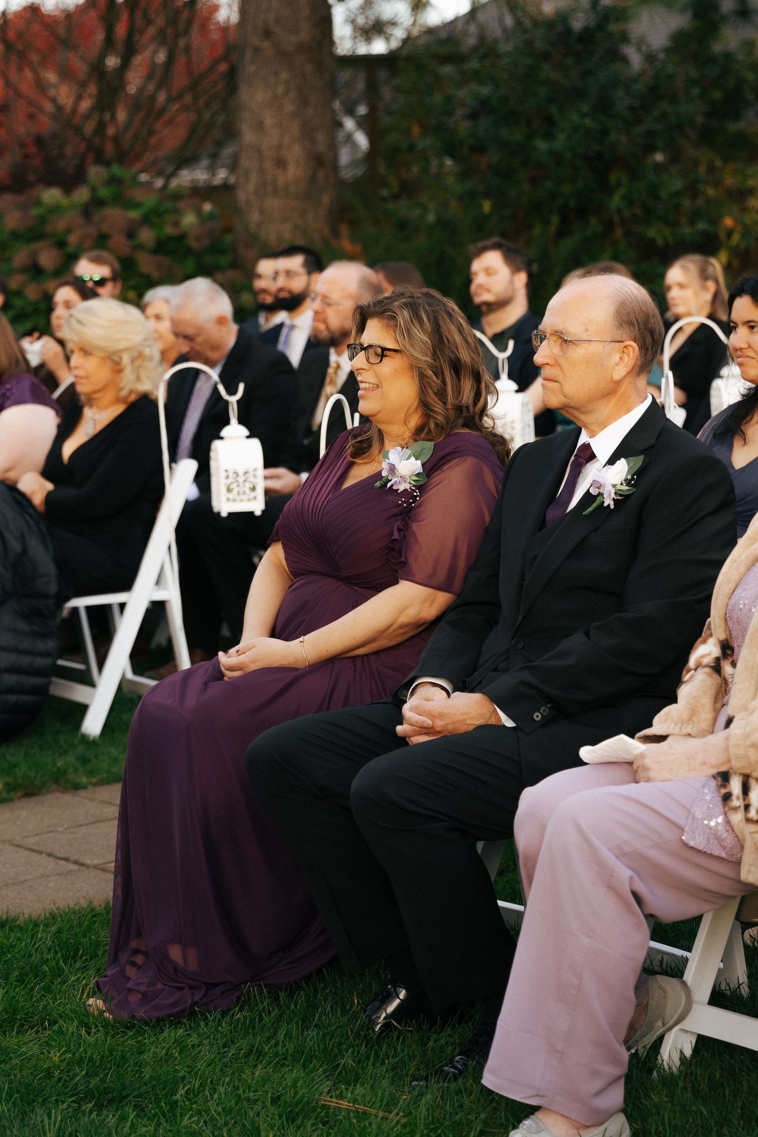 Wedding guests seated in white chairs outdoors watching ceremony in purple and formal attire.