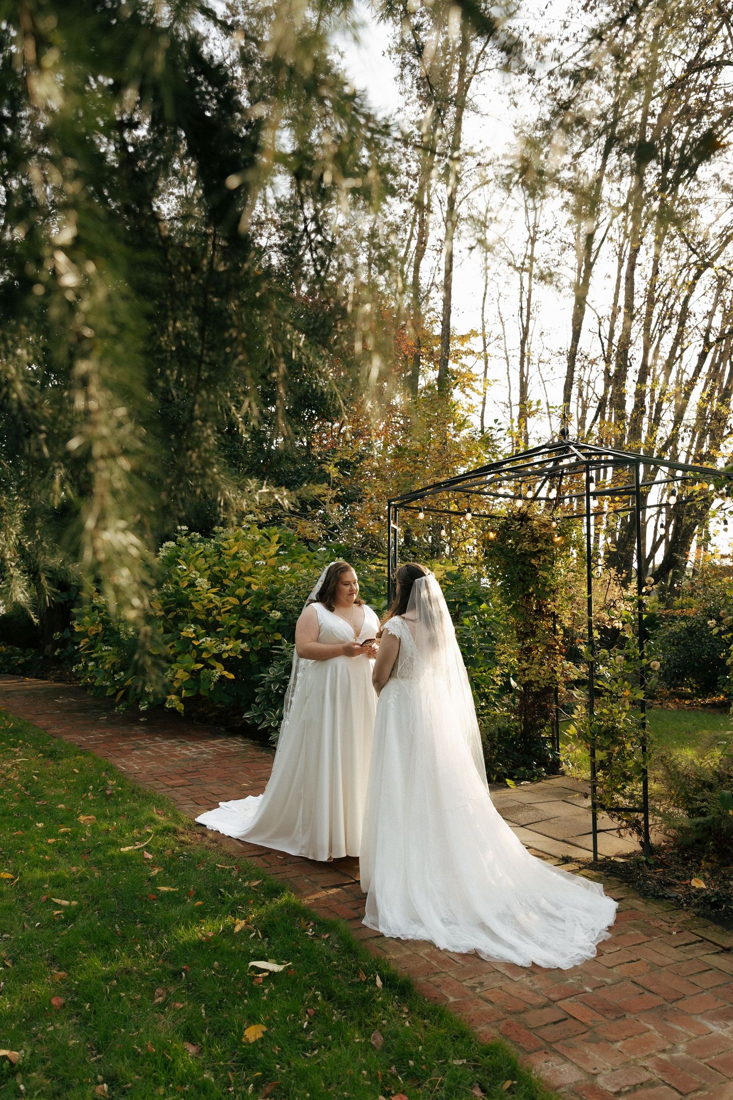 A bride in a flowing white dress stands on a brick path under a garden arch surrounded by autumn foliage.