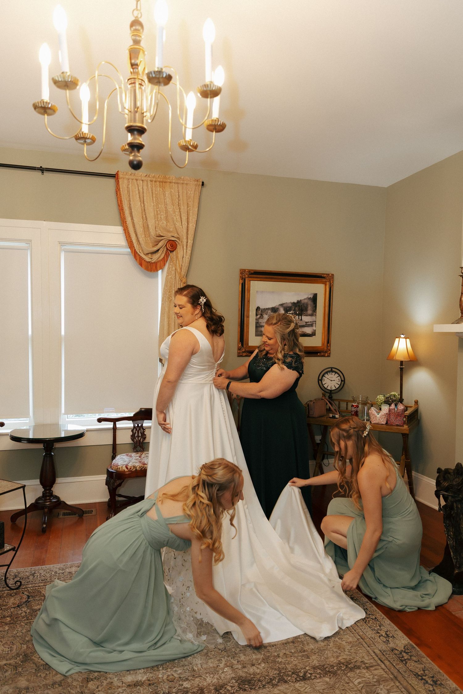Bridesmaids in sage green dresses help adjust a white wedding gown in an elegant living room setting.