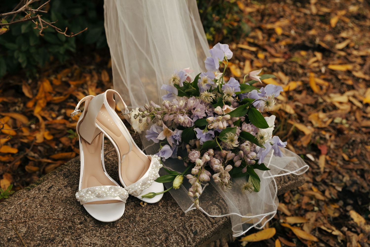 White bridal shoes and delicate floral bouquet with purple flowers displayed on autumn leaves.