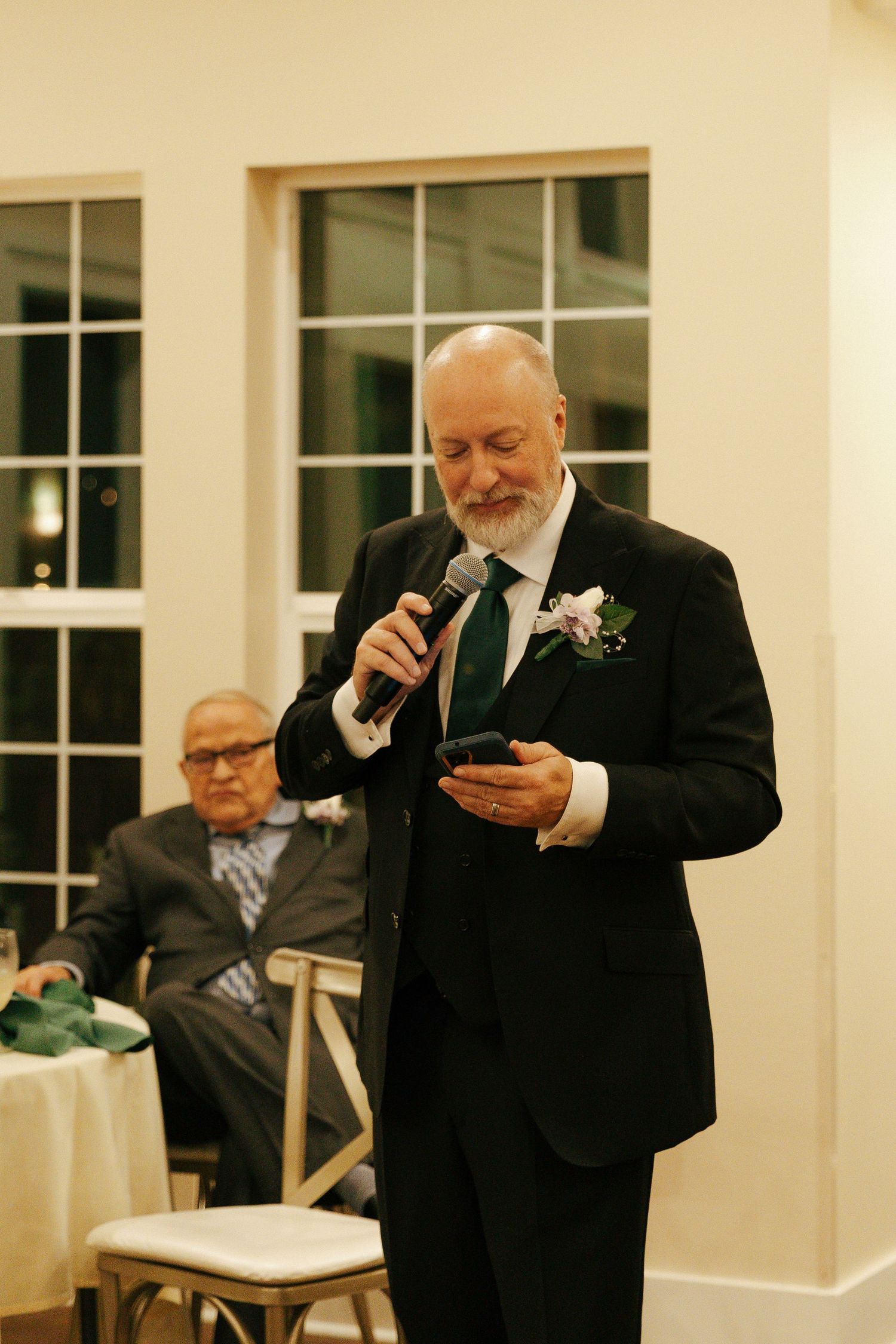 Man in formal black suit reading from phone during what appears to be a wedding speech at an indoor venue.