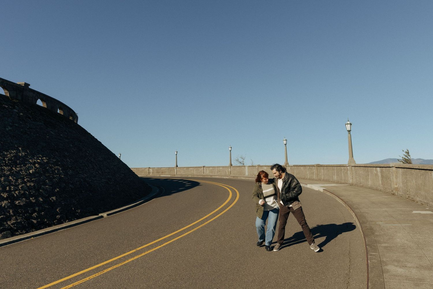 Couple standing close together on curved coastal road with blue sky.