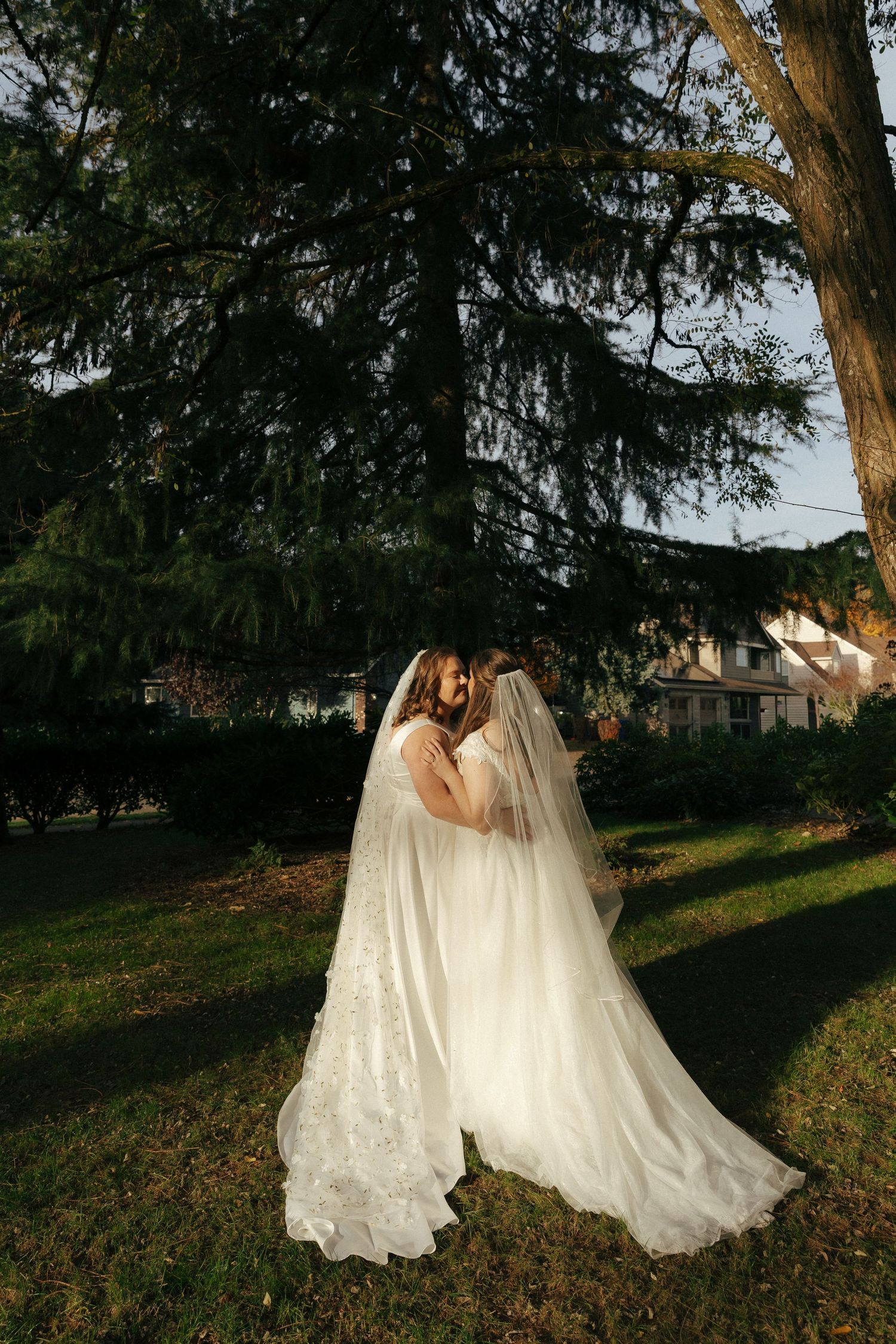 Two figures in flowing white wedding gowns dance together under tall trees in a romantic outdoor setting at sunset.