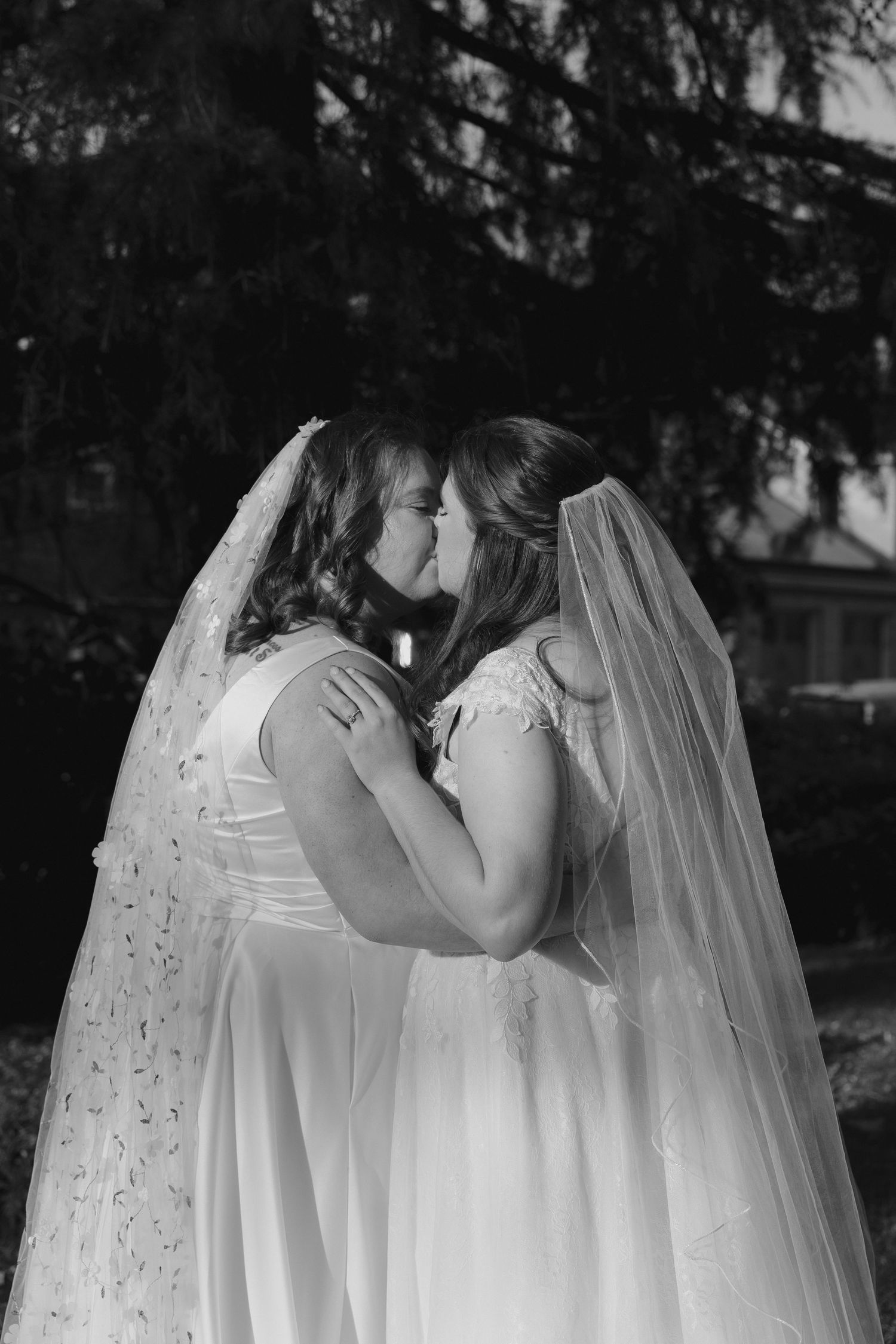 Two people in white wedding dresses share a tender kiss while their veils flow behind them in a black and white photo.