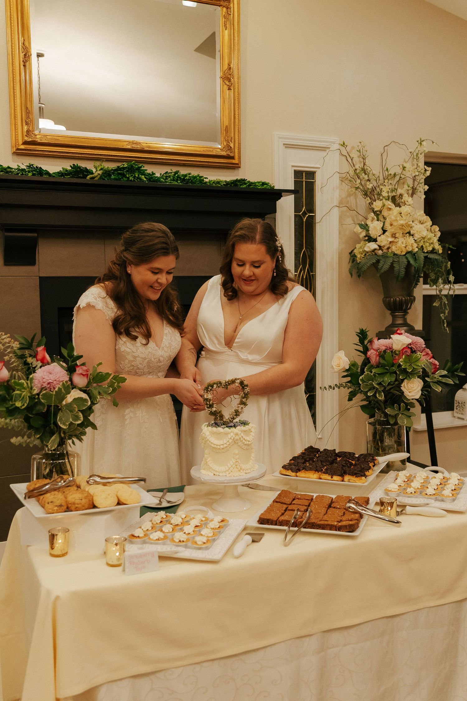 Two people in white dresses cutting desserts at an elegant reception table with floral arrangements.