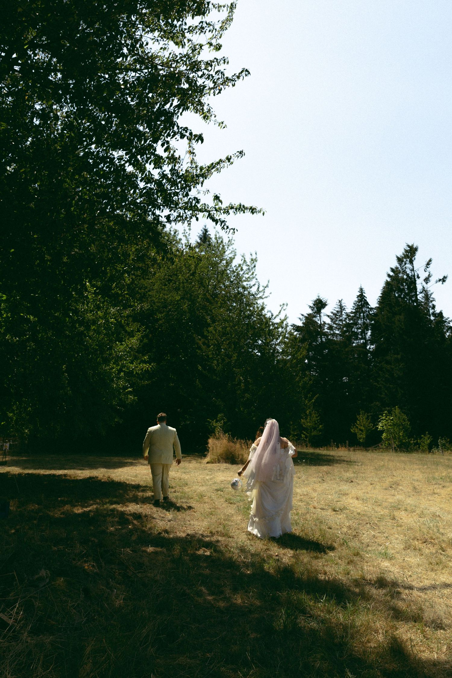 A bride in a white gown and veil walks with a groom in a tan suit through a sunlit meadow surrounded by lush green trees.