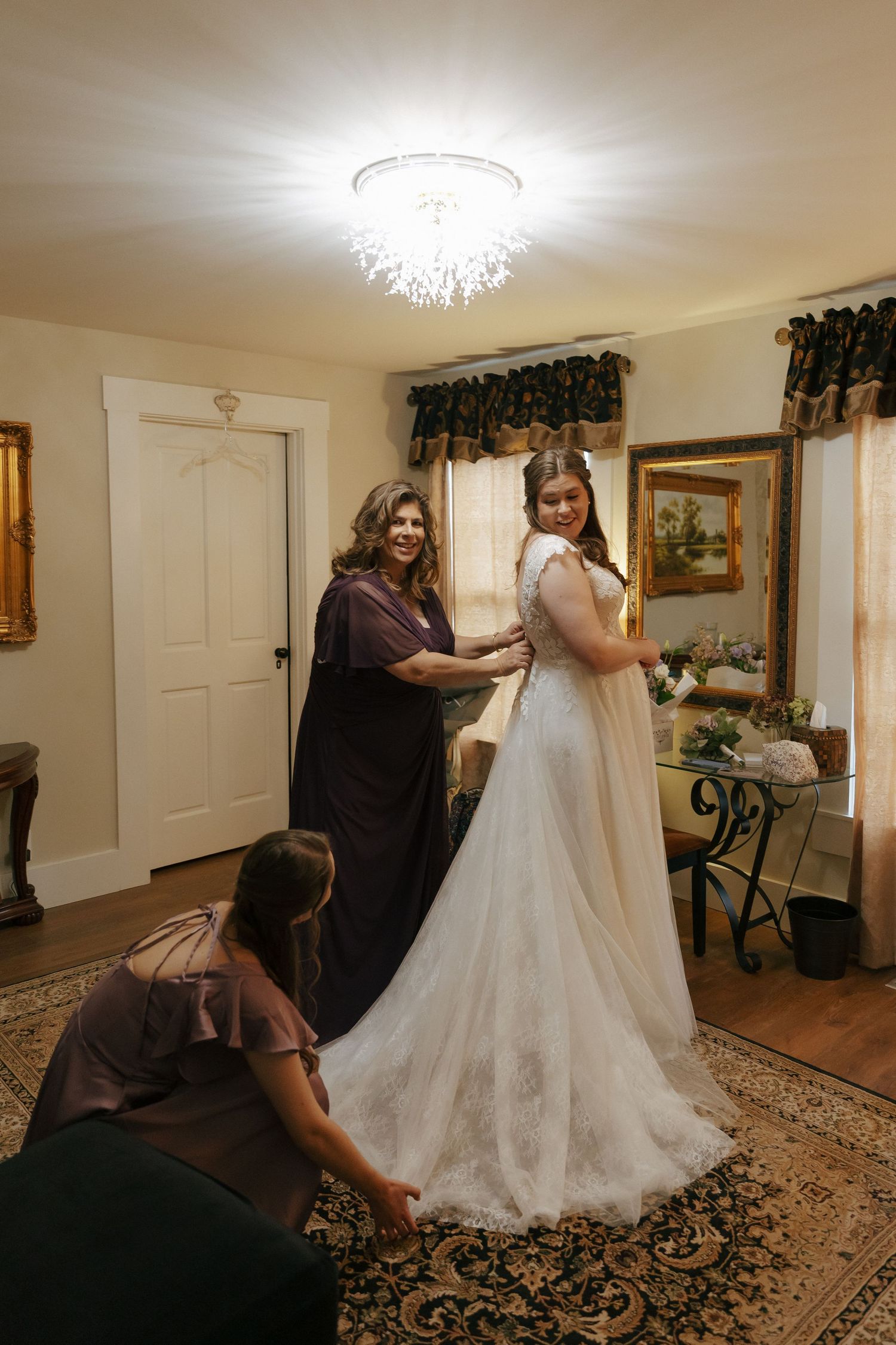 Wedding preparations in a bridal suite showing the final dress adjustments and fittings on an ornate patterned rug.