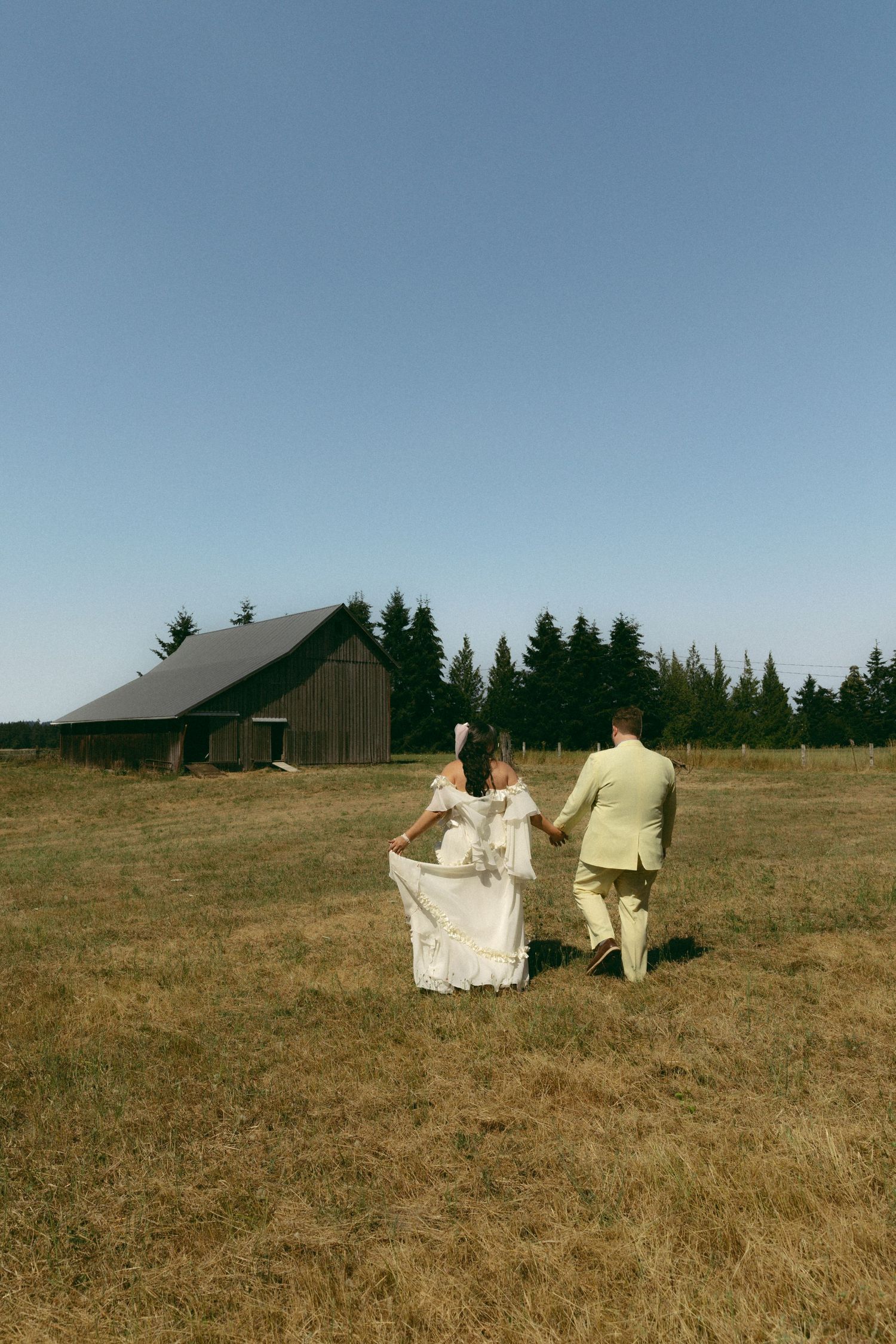 Bride and groom walking hand in hand across a golden field toward a rustic barn, surrounded by tall evergreen trees.