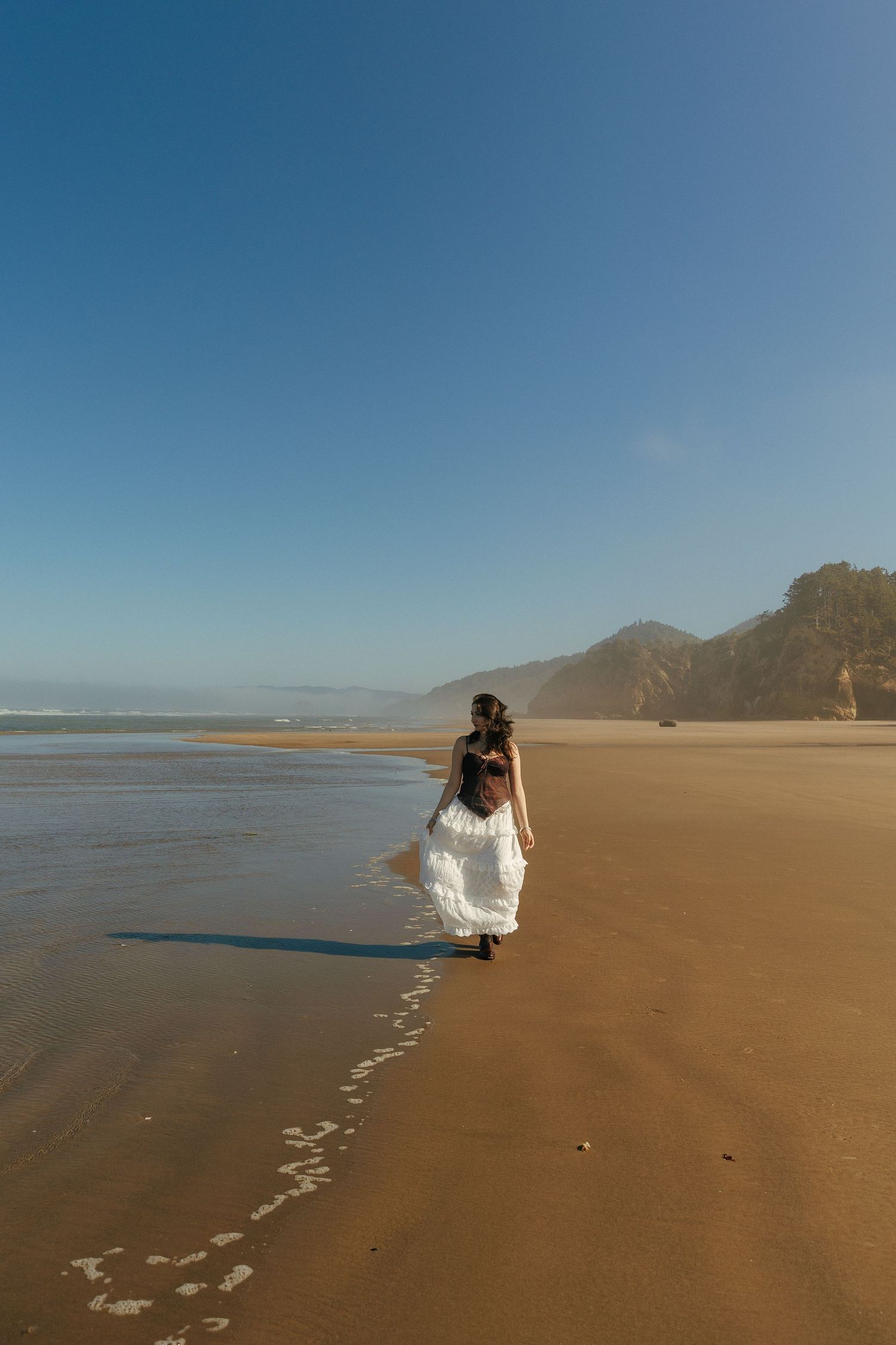 Senior photos in white dress walking on the beach leaving footprints in sand.
