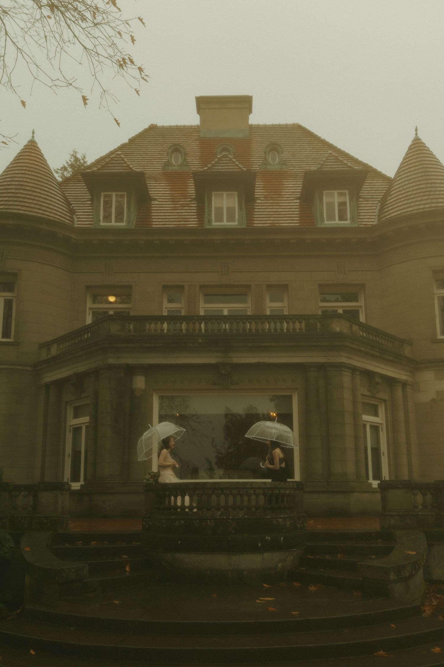 Historic mansion in foggy weather with couple holding umbrellas on balcony.