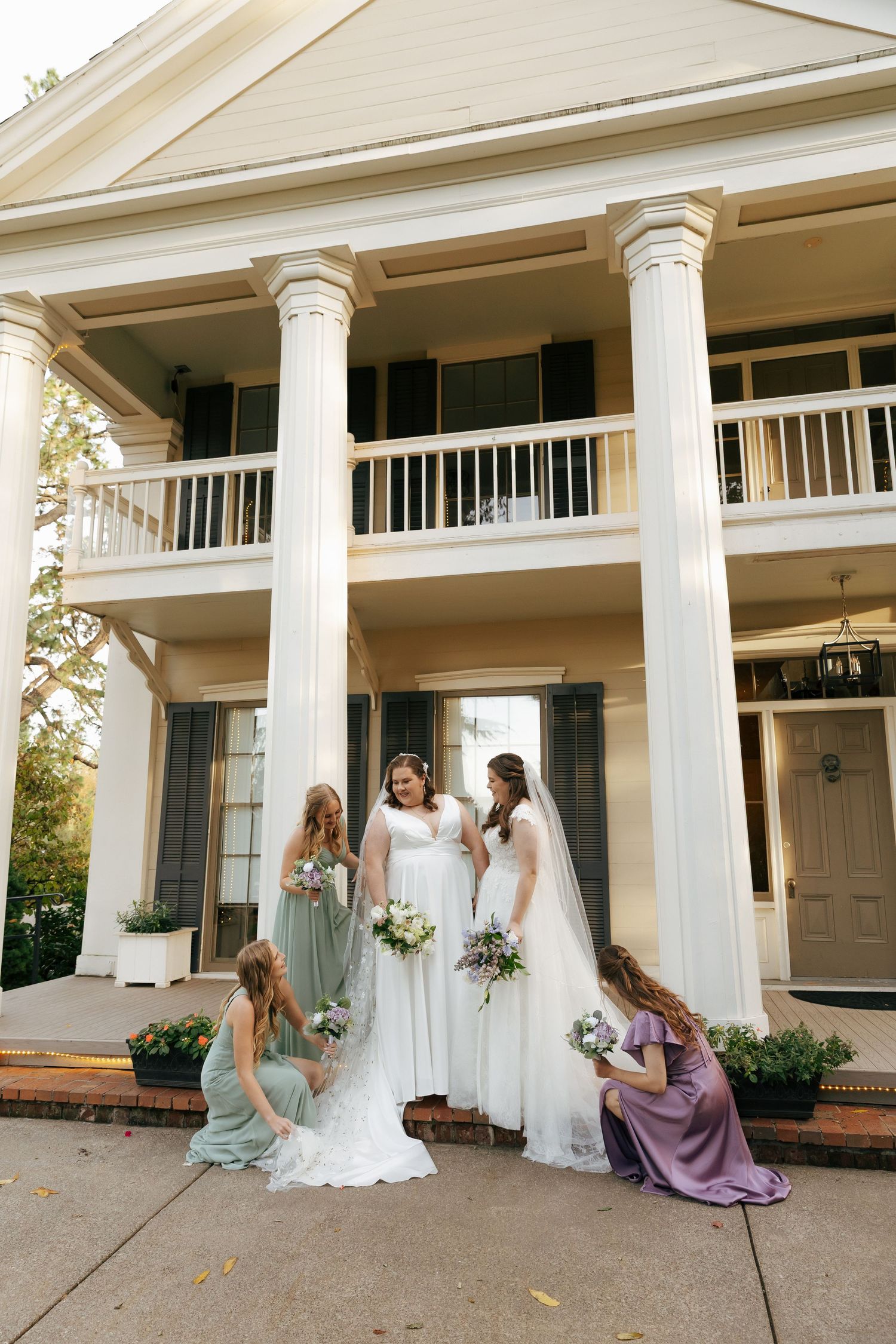 A bridal party in pastel dresses gathers on a white-columned porch during a wedding photoshoot.