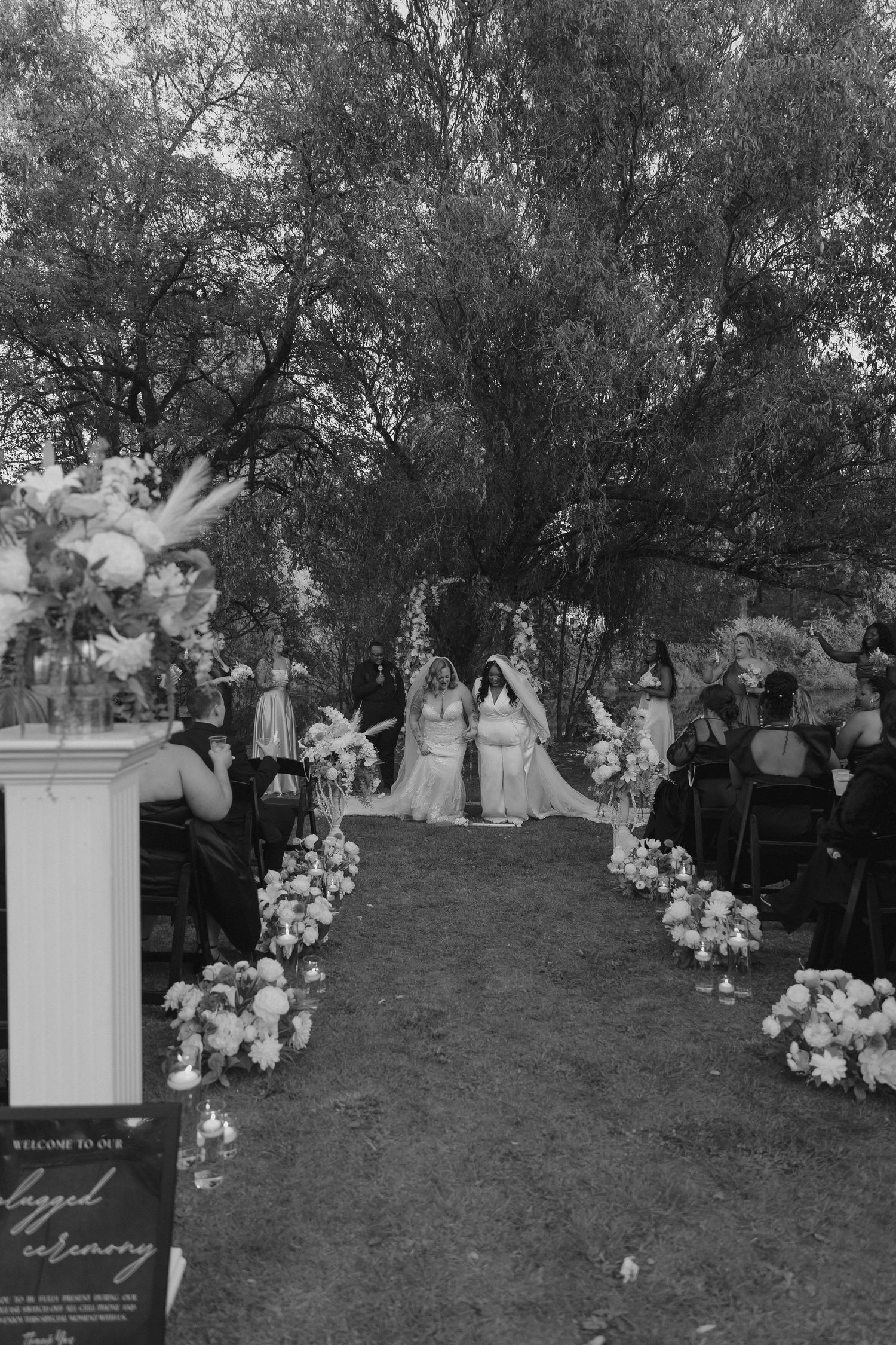 A black and white outdoor wedding ceremony with floral arrangements lining the aisle under tall trees.