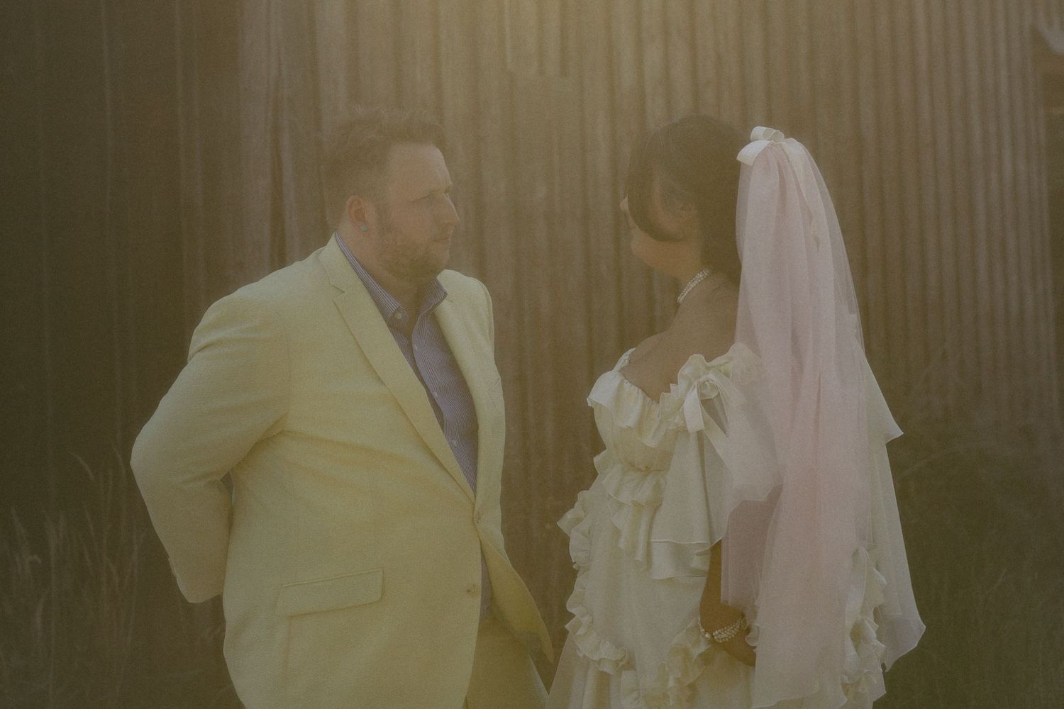 A groom in a yellow suit and bride in a white ruffled dress with veil share a moment near a rustic barn.