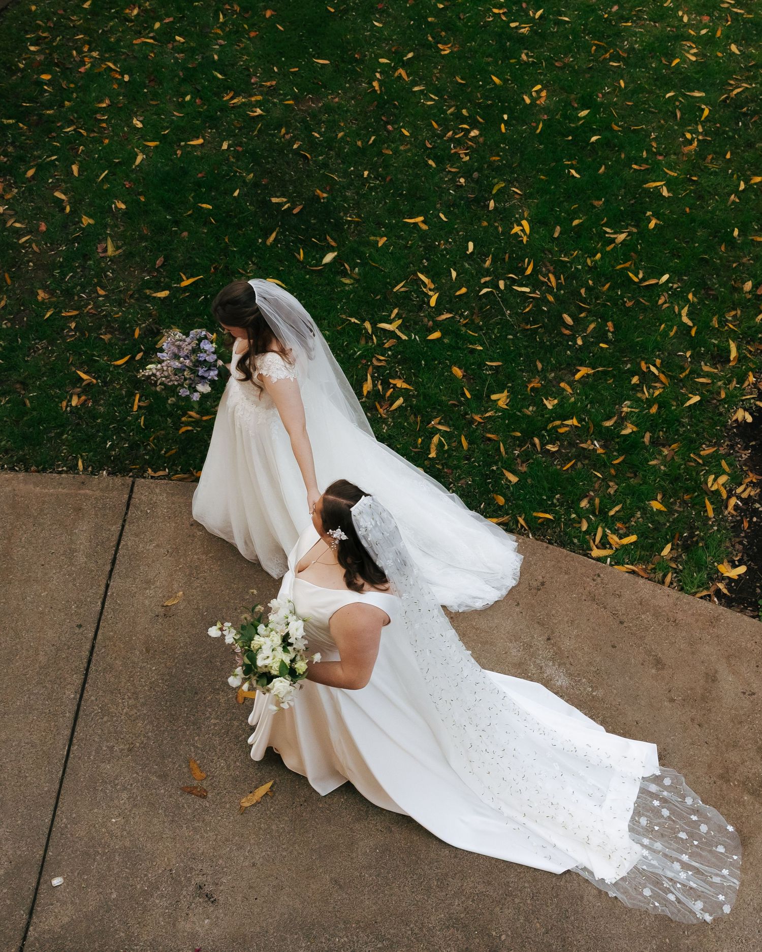 Two brides in flowing white dresses walk along a path with scattered autumn leaves.