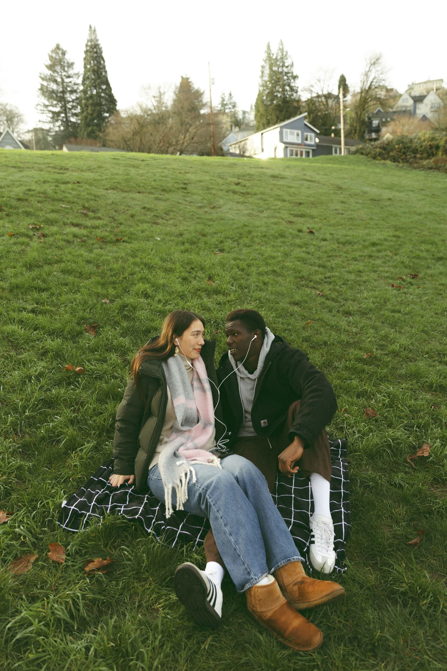 A couple sits together on a plaid blanket in a grassy field on an autumn day.