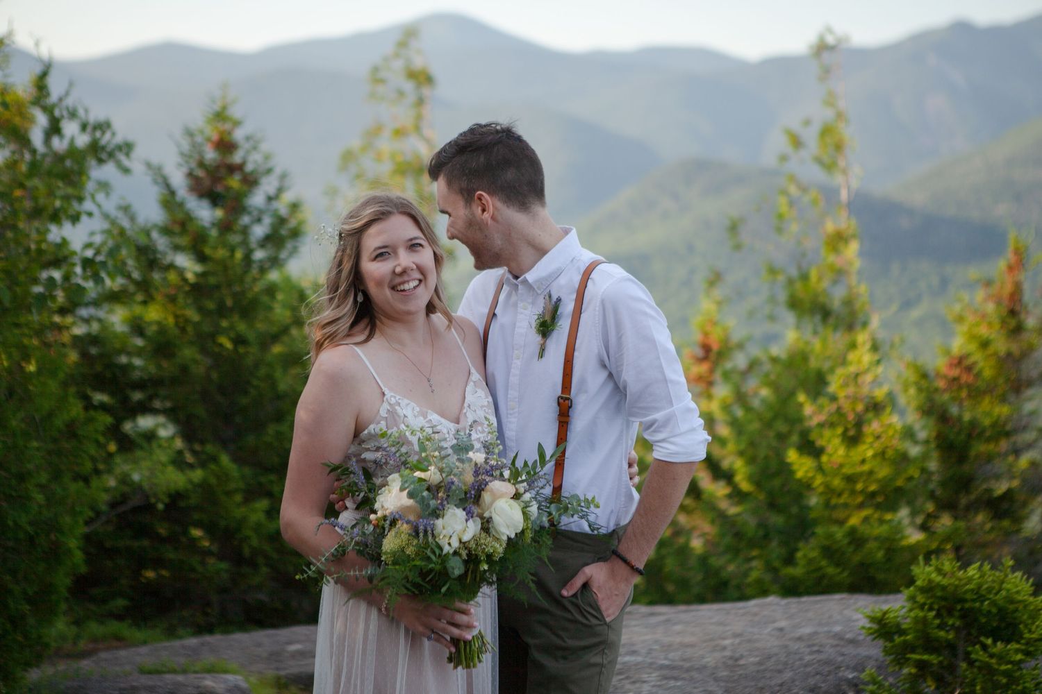 Couple smiling together at mountain overlook, bride holding wildflower bouquet, groom in suspenders, lush green peaks behind.